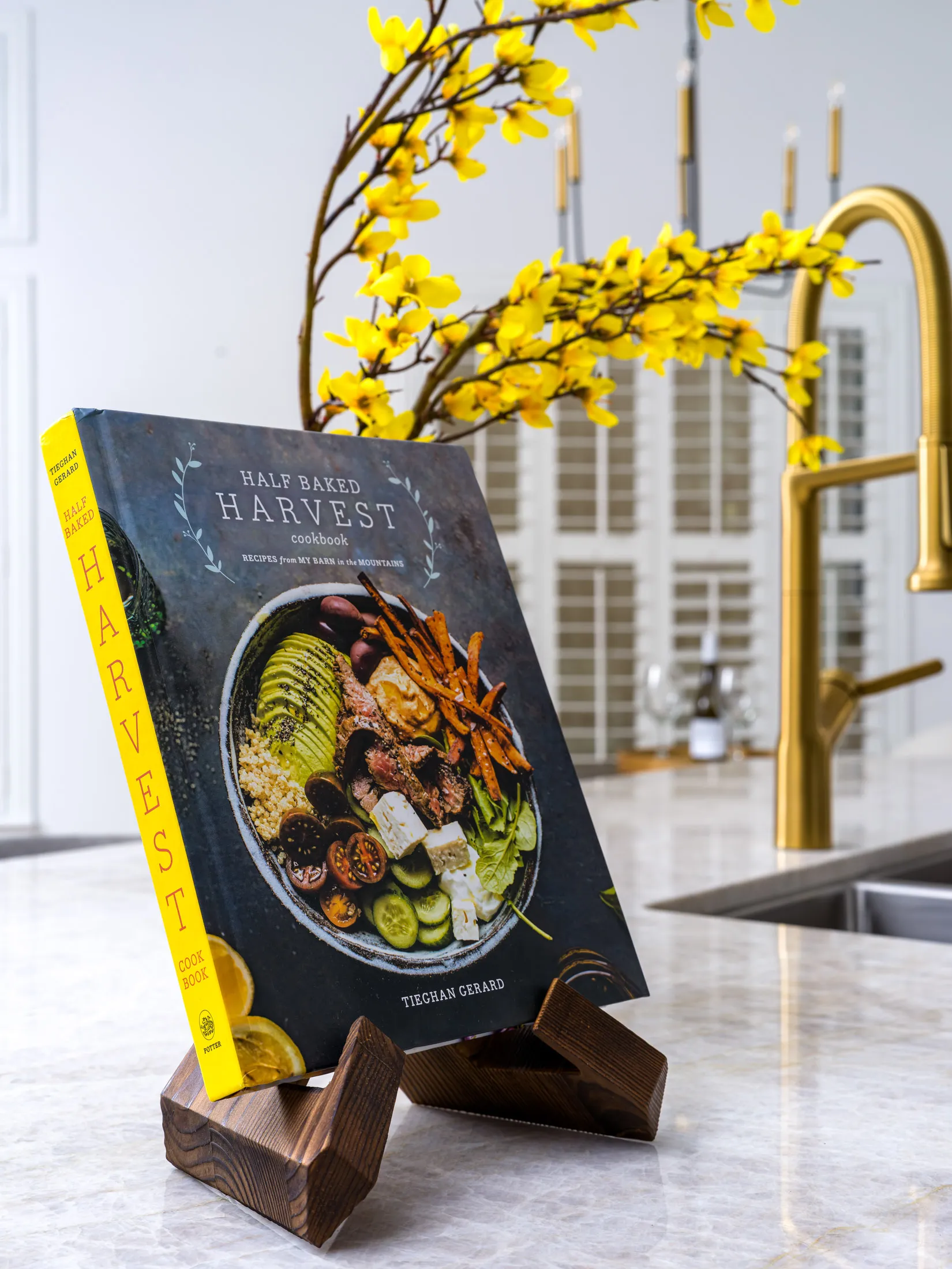 A hardcover cookbook on a wooden stand, with bright yellow blossoms and a gold faucet in the background.