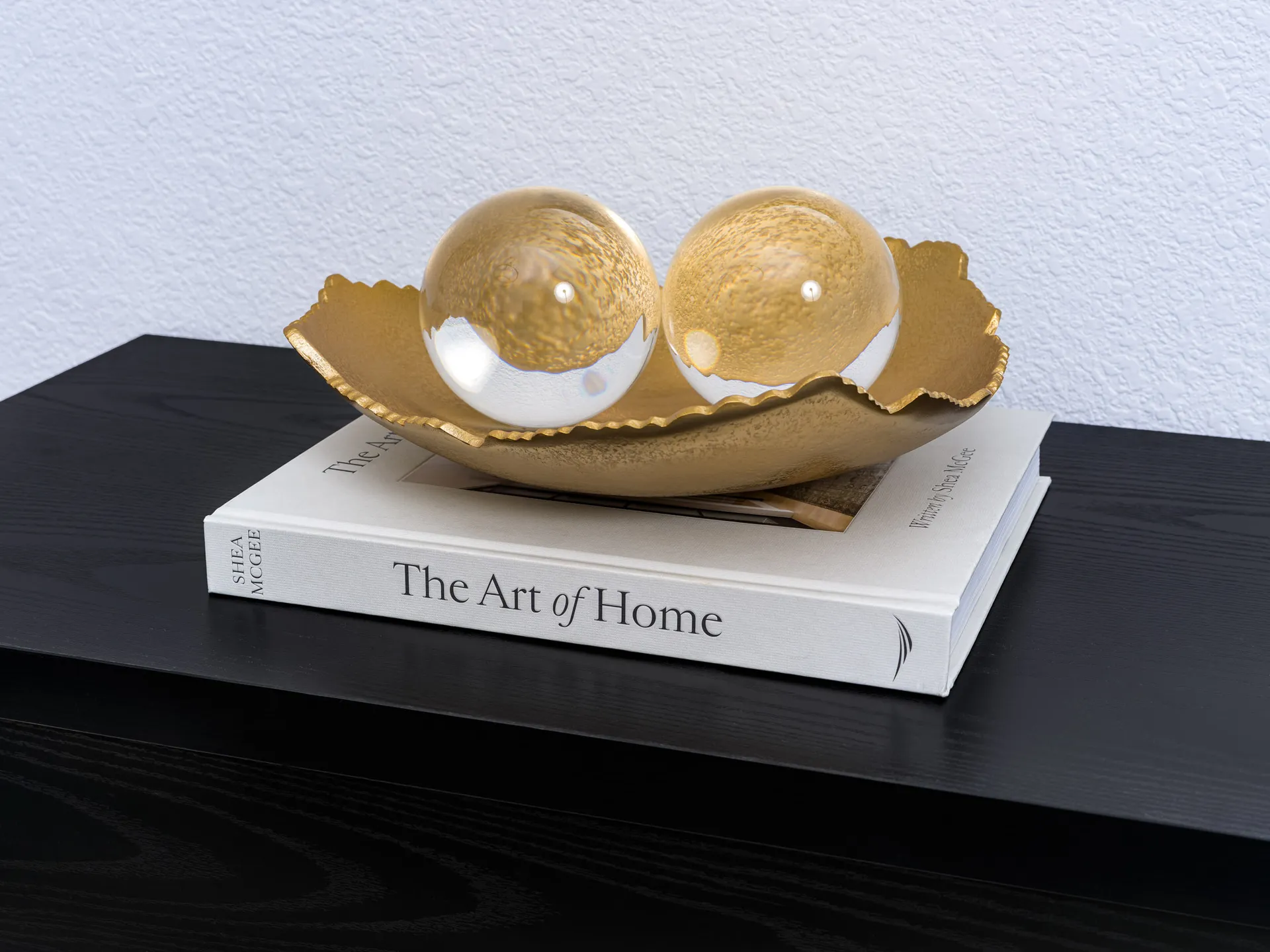 A gold tray with scalloped edges holding two clear glass spheres, resting on a hardcover book atop a black console.