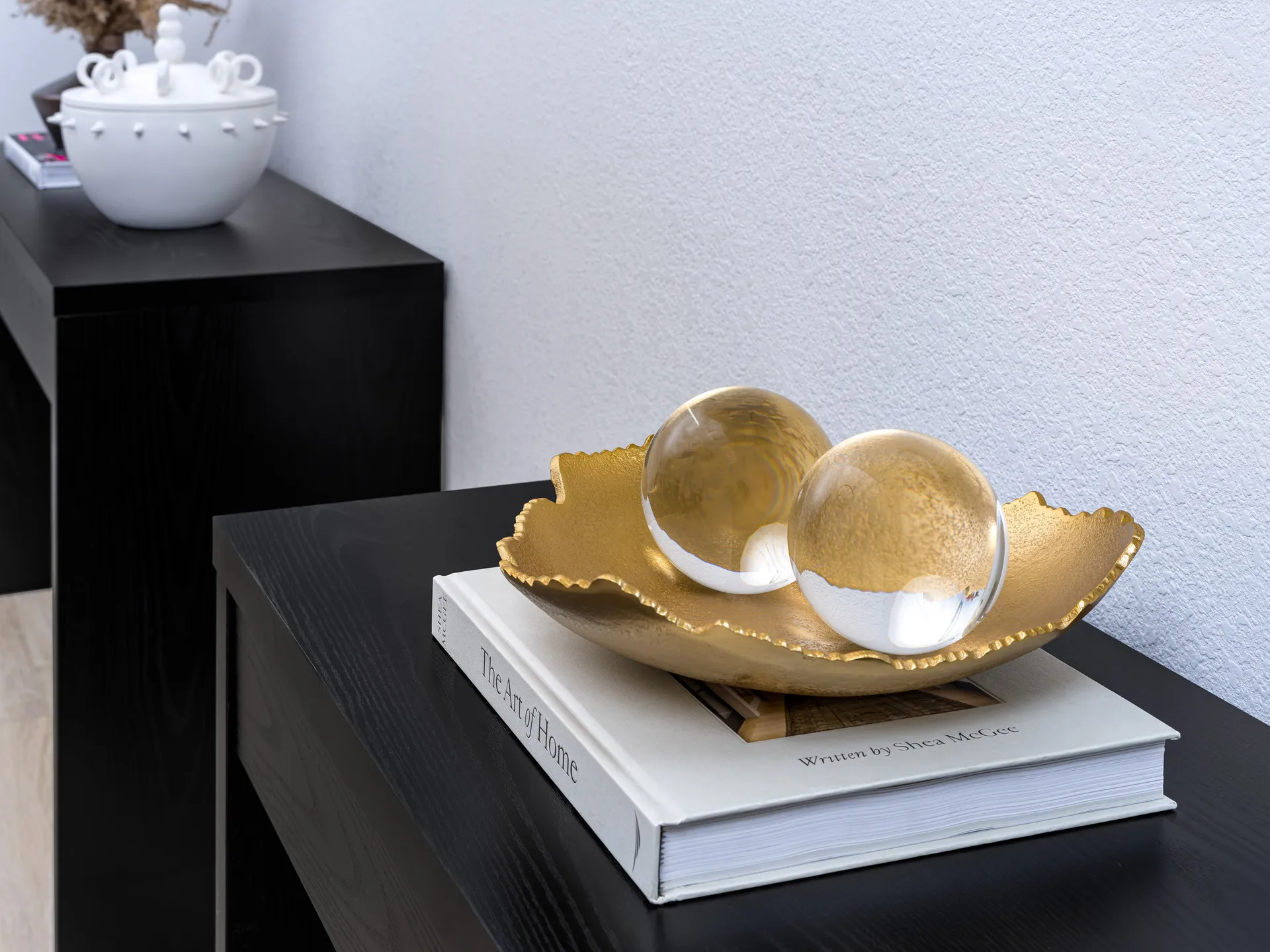 A gold tray with ruffled edges holding two clear glass spheres, resting on a hardcover book on a black console.
