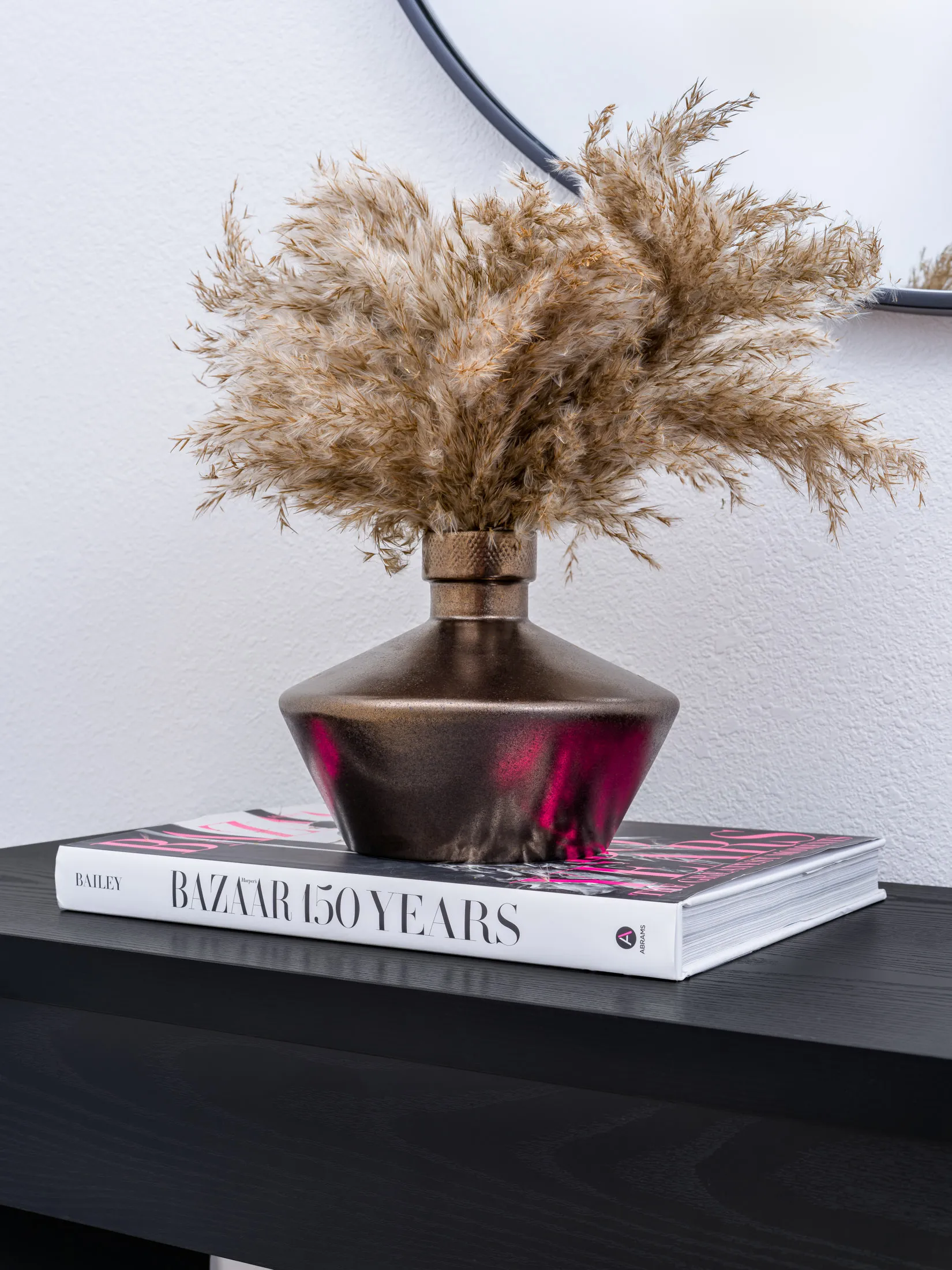 A bronze-toned vase filled with dried pampas grass, resting on a fashion book atop a black console.