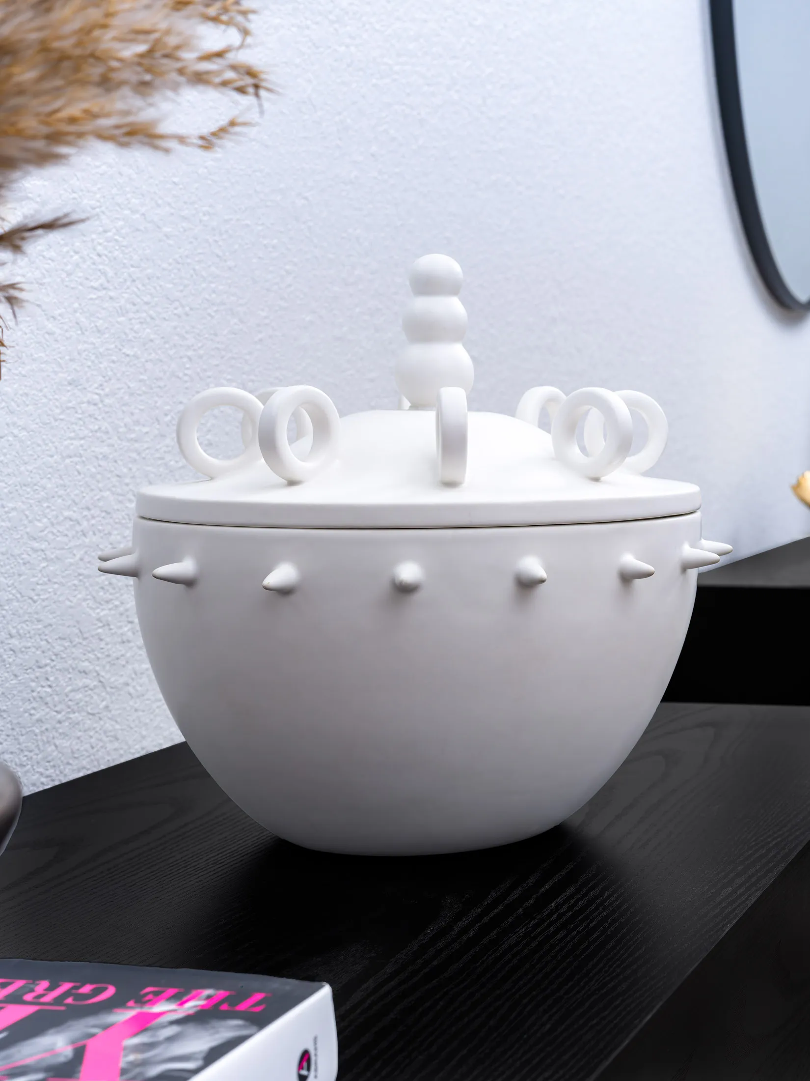 A white ceramic container with looped handles and small spikes on its lid, displayed on a black console.