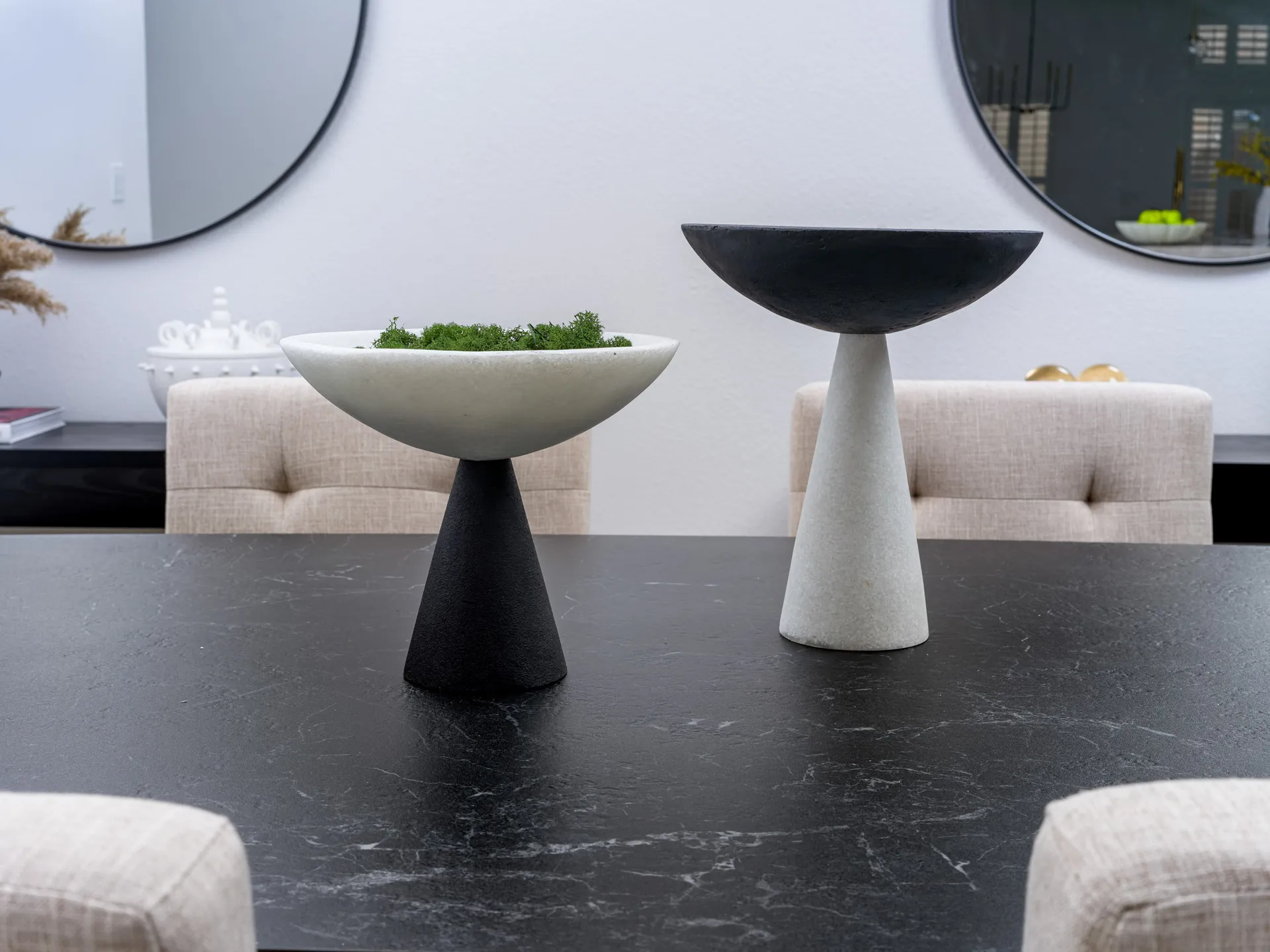 Two modern pedestal bowls in black and white on a dark dining table with upholstered chairs and round mirrors in the background.