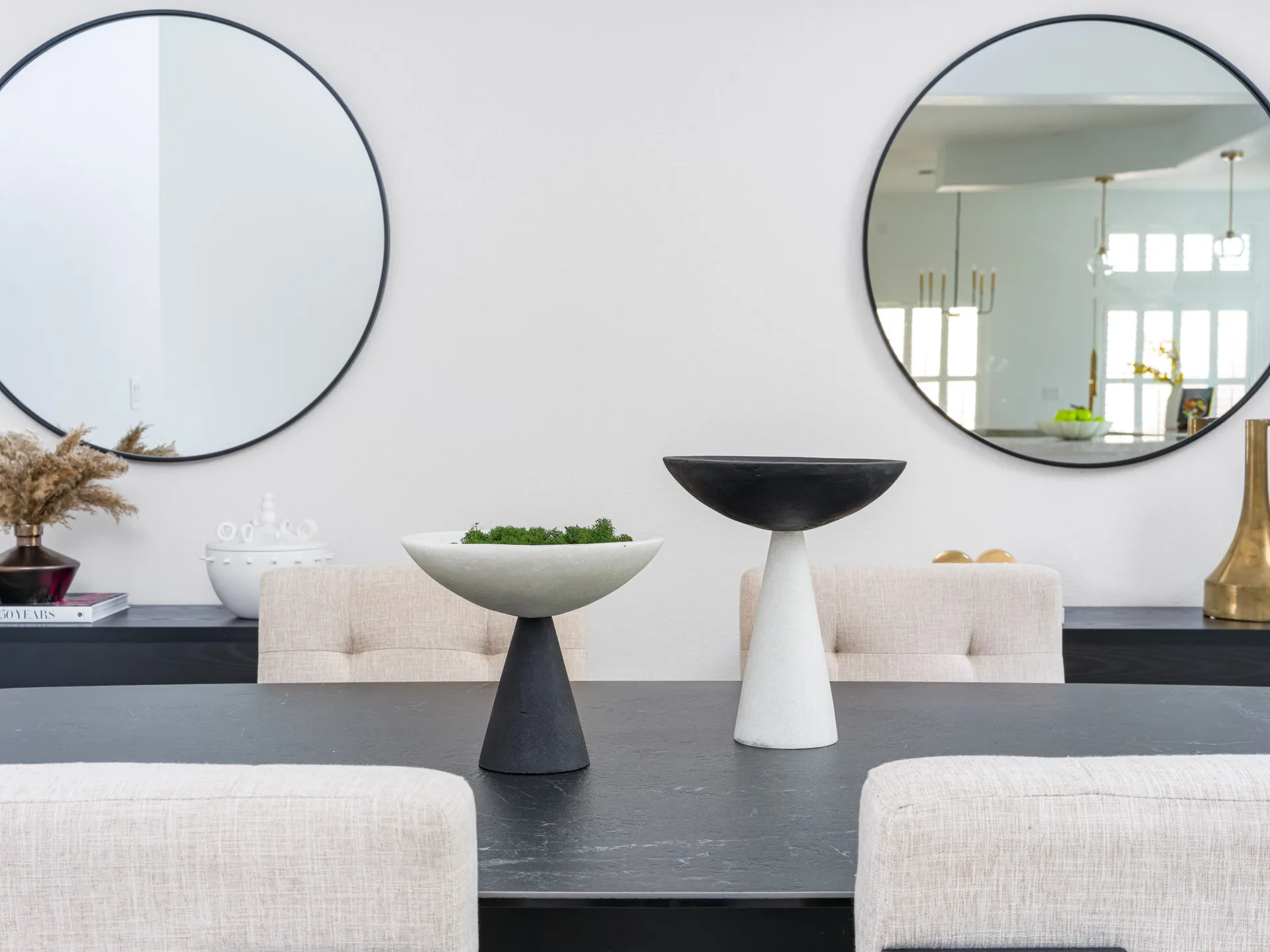 A dark dining table with neutral chairs, featuring two sculptural pedestal bowls and round wall mirrors.