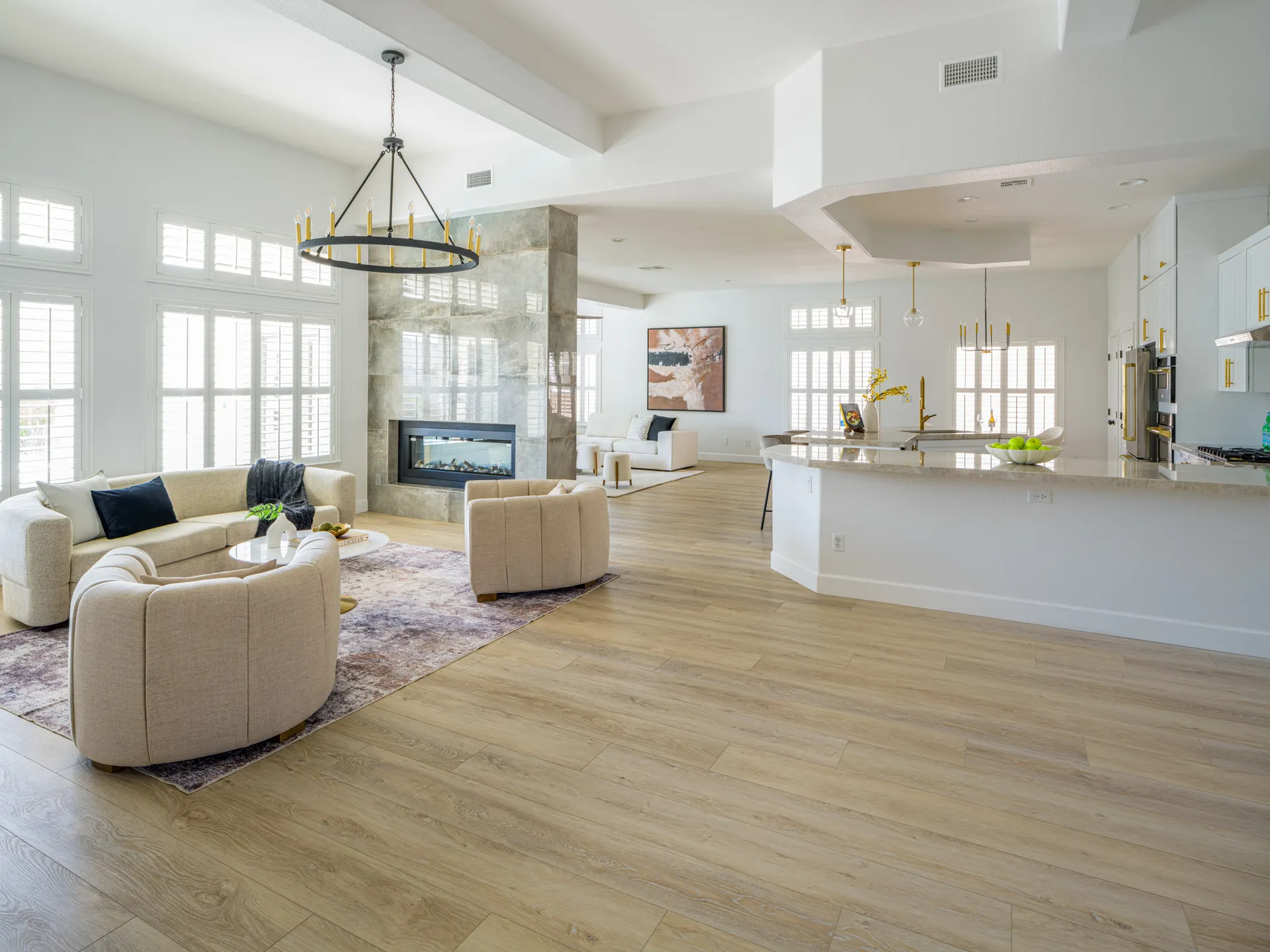 A large open-concept living space with tall windows, a modern fireplace, neutral sofas, and a kitchen island in the background.