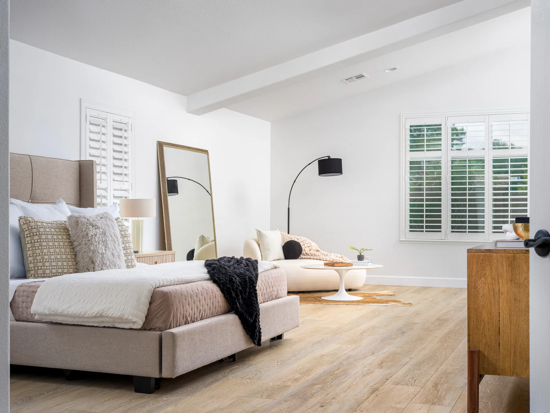 A spacious bedroom with a neutral upholstered bed, large floor mirror, and cozy sitting area featuring a round coffee table.