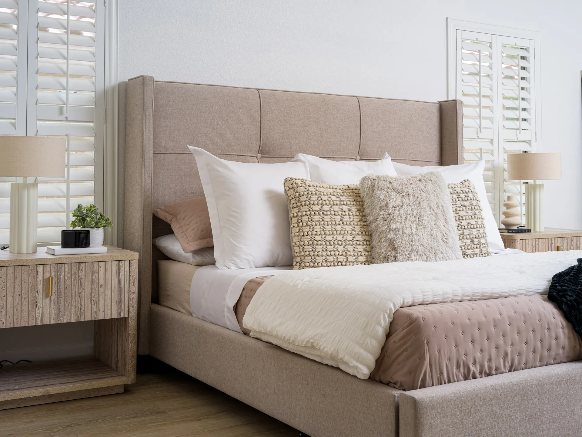 A beige upholstered bed with layered pillows and matching nightstands beside shuttered windows.