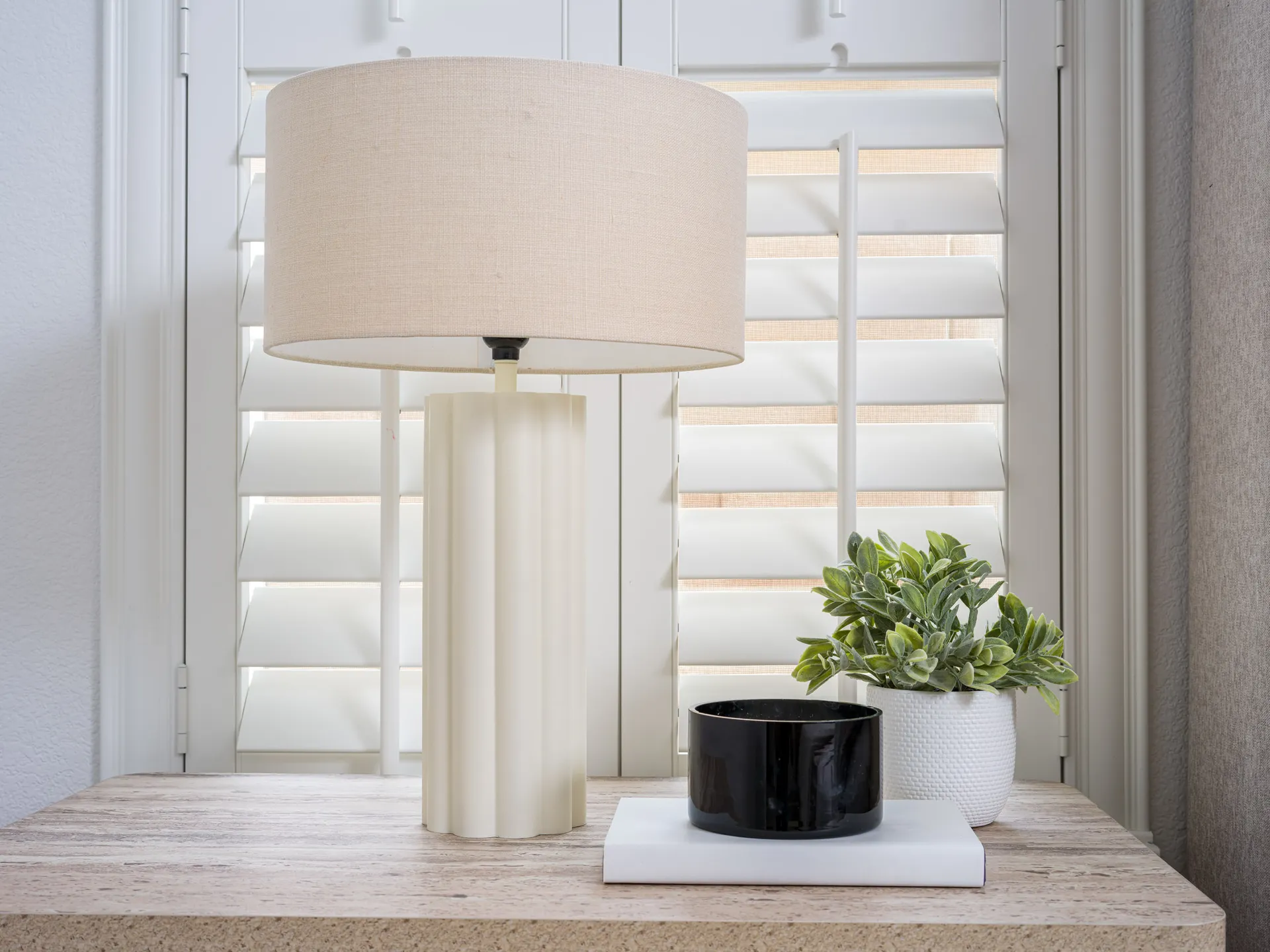 A cream fluted lamp on a nightstand with a black bowl and a white planter of green leaves, set against white shutters.