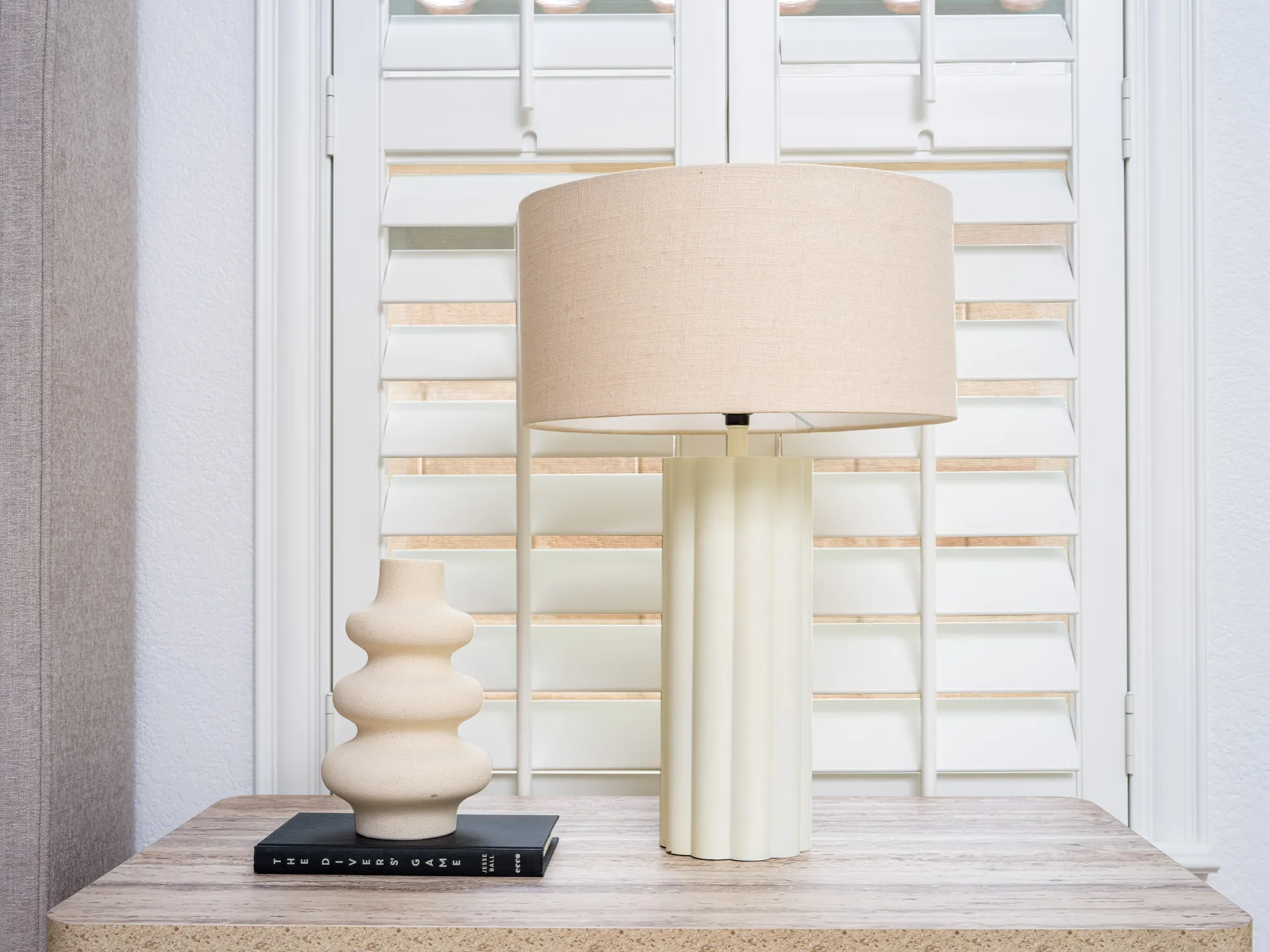 A fluted cream lamp and a beige sculptural vase on a nightstand, in front of white shutters.