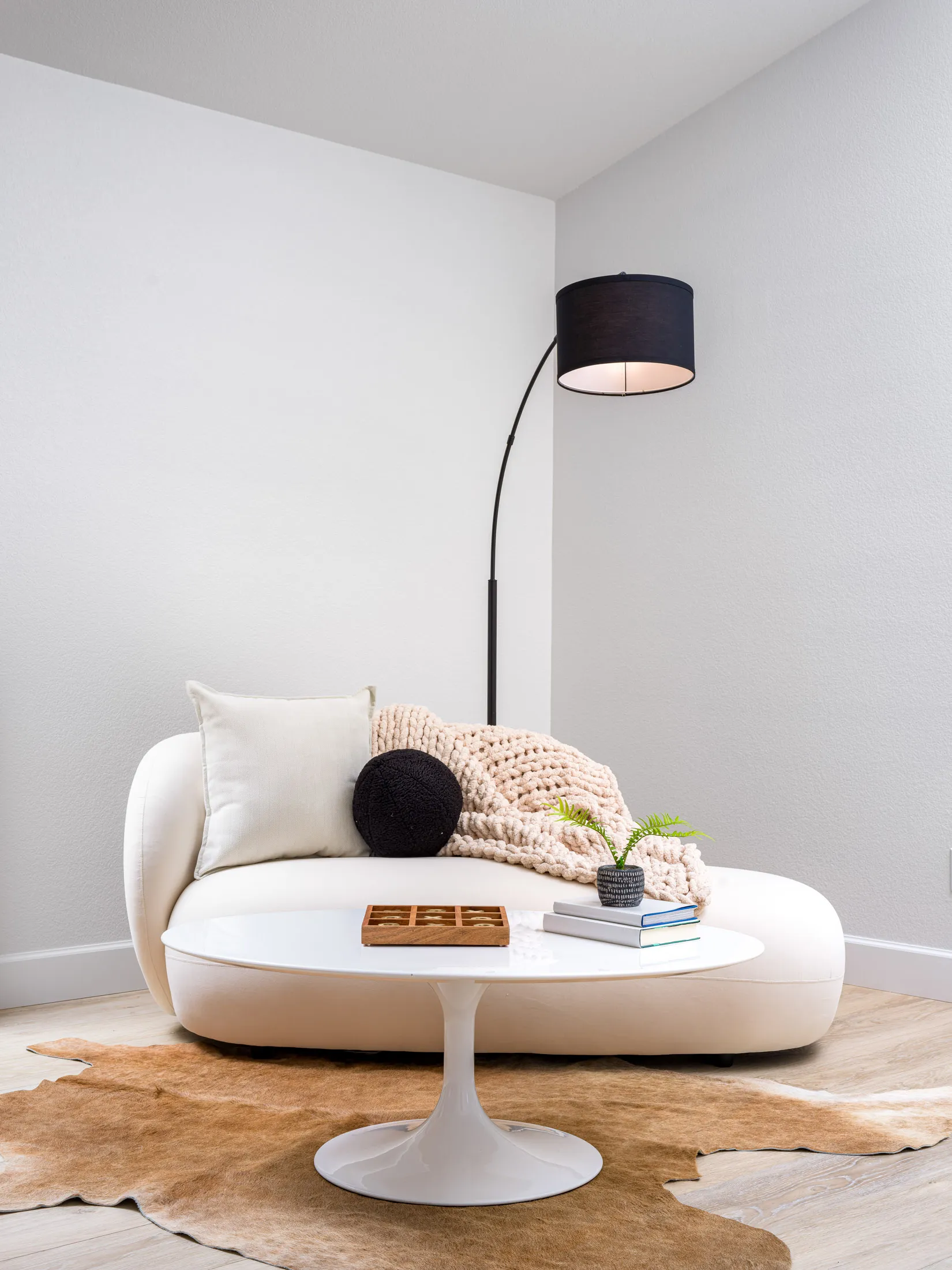 A curved white sofa with a chunky knit throw, a black arc floor lamp, and a round white coffee table atop a cowhide rug.