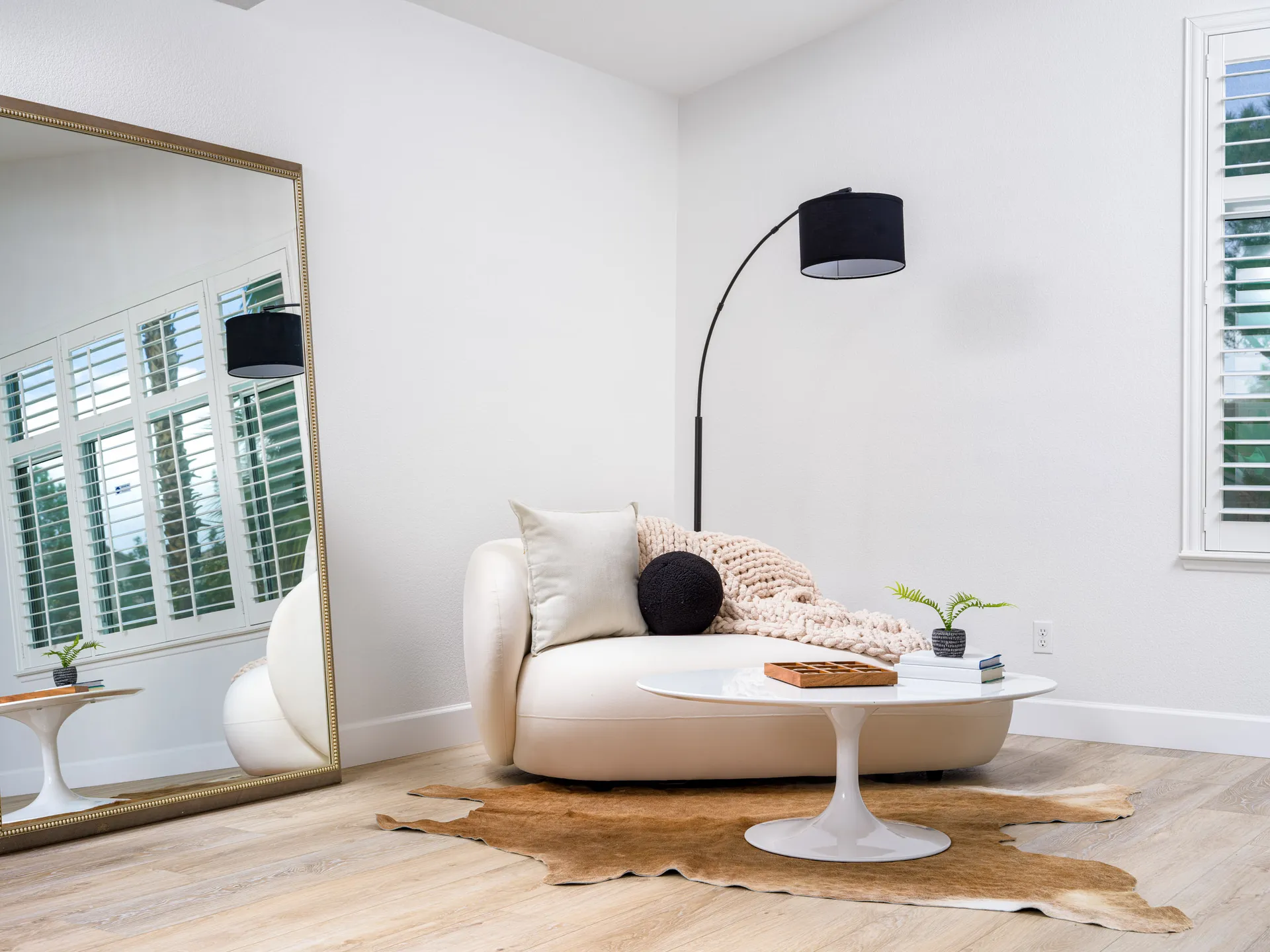 A cozy corner with a curved white sofa, a tall black floor lamp, a large mirror, and a white coffee table topped with a wooden tic-tac-toe set.