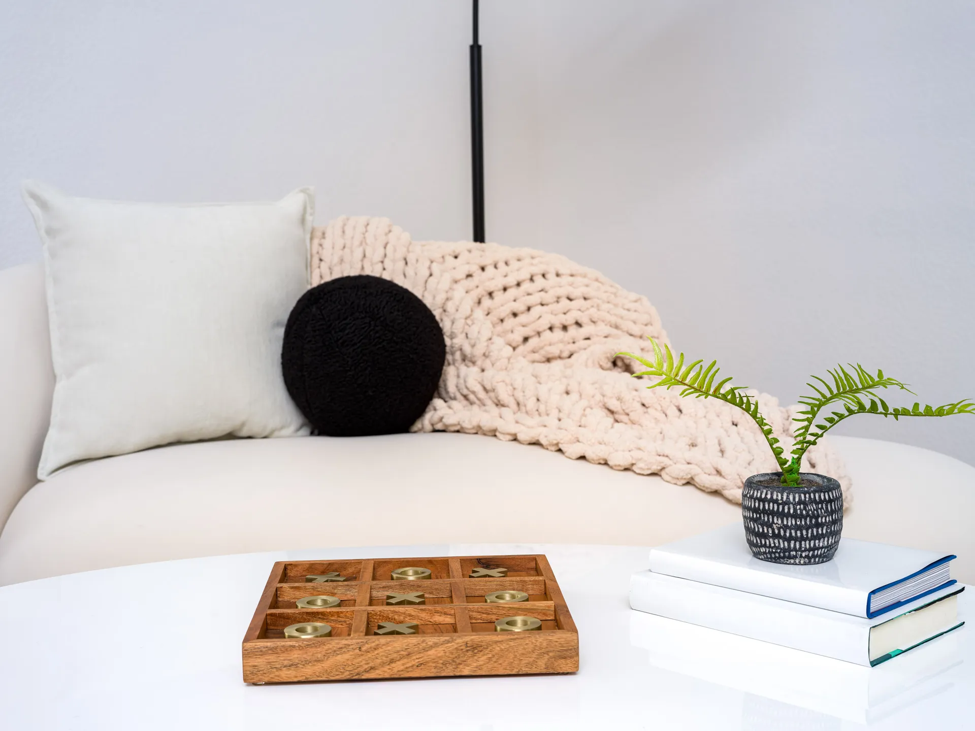 A white sofa with plush pillows and a chunky knit throw, with a coffee table displaying a wooden tic-tac-toe set and a small potted plant on stacked books.