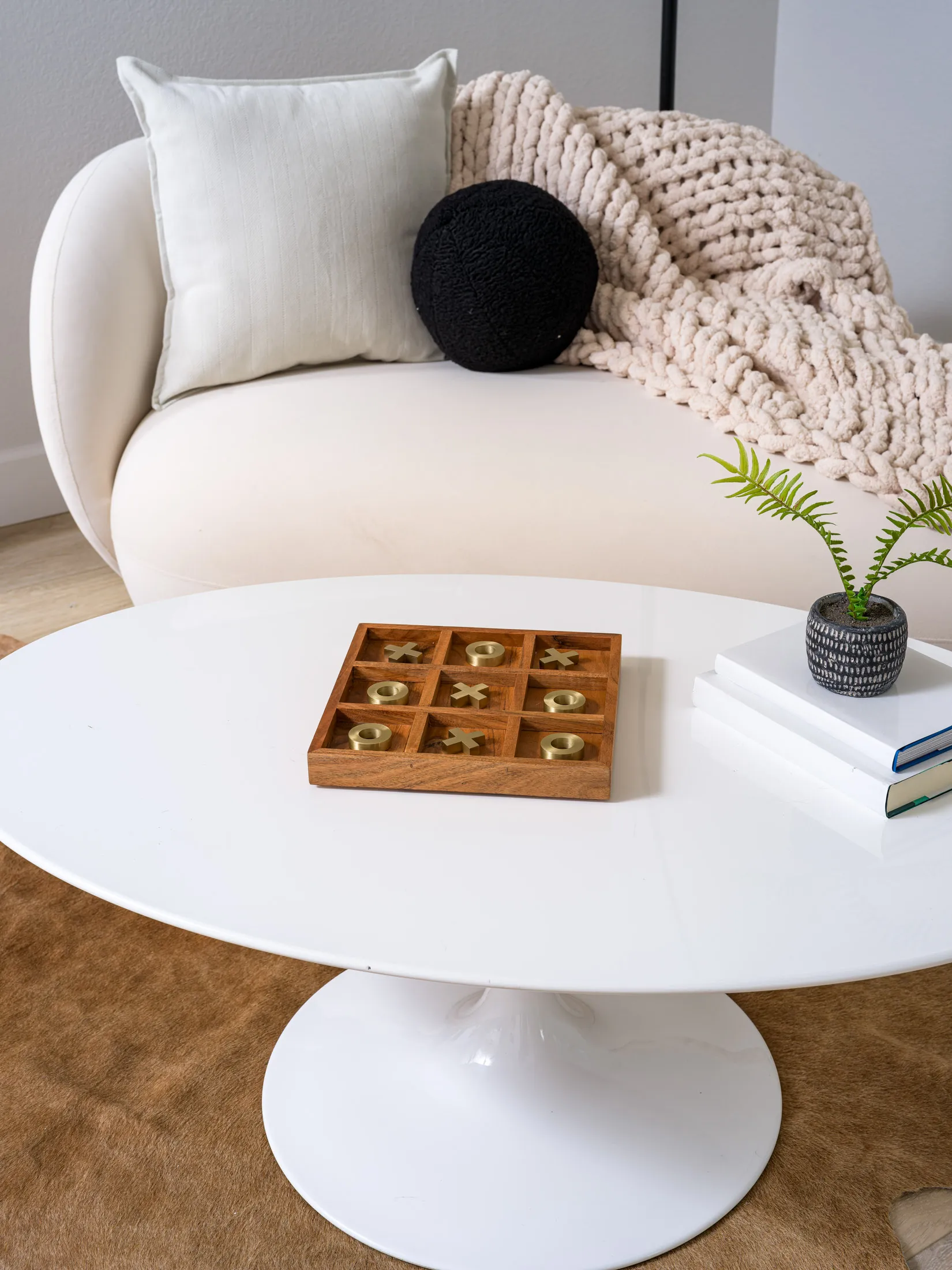 A round white coffee table displaying a wooden tic-tac-toe set with gold pieces, a small potted plant, and stacked books in front of a plush white sofa.