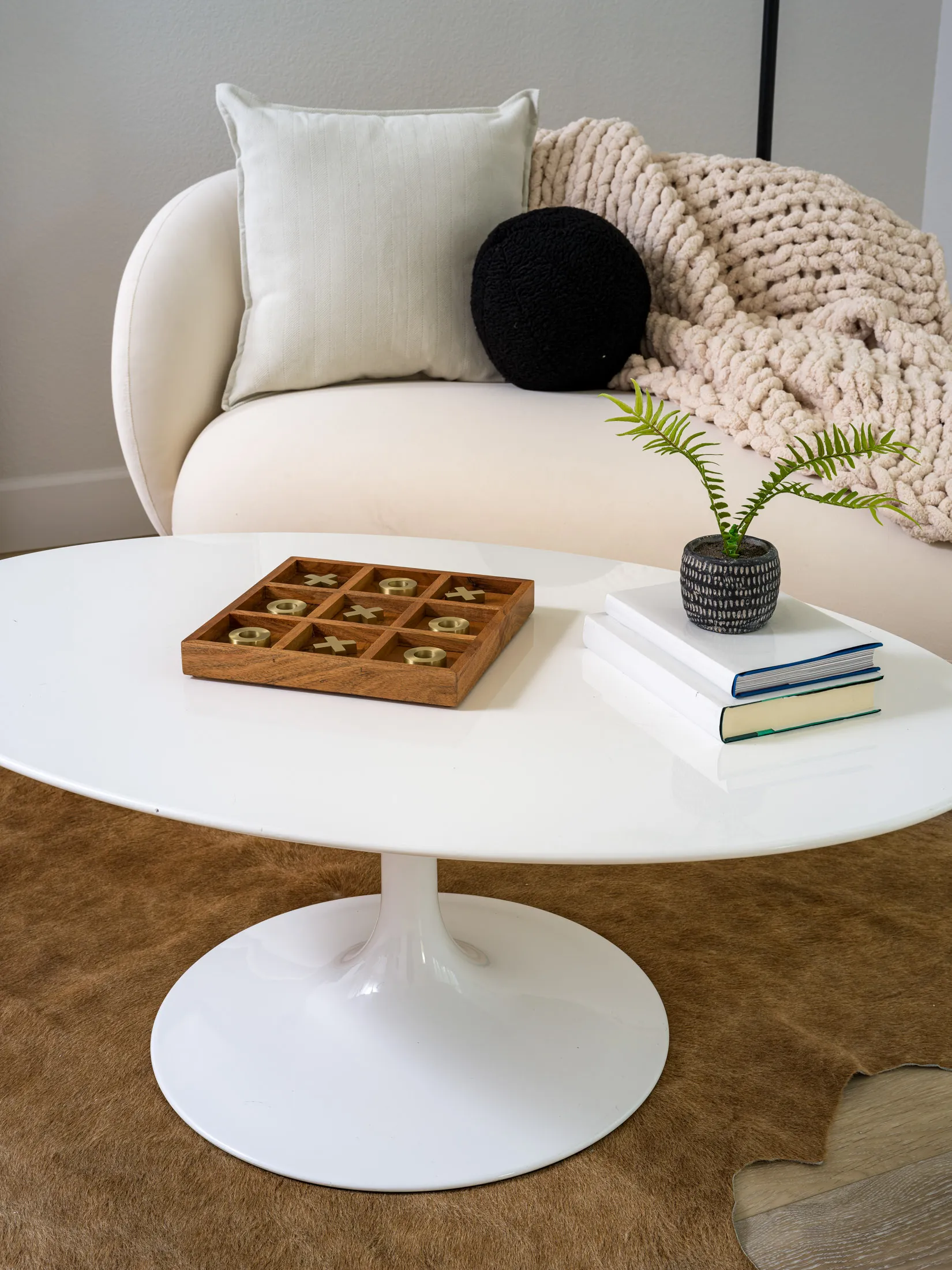 A round white coffee table with a wooden tic-tac-toe game and gold pieces, next to a small potted plant and stacked books.