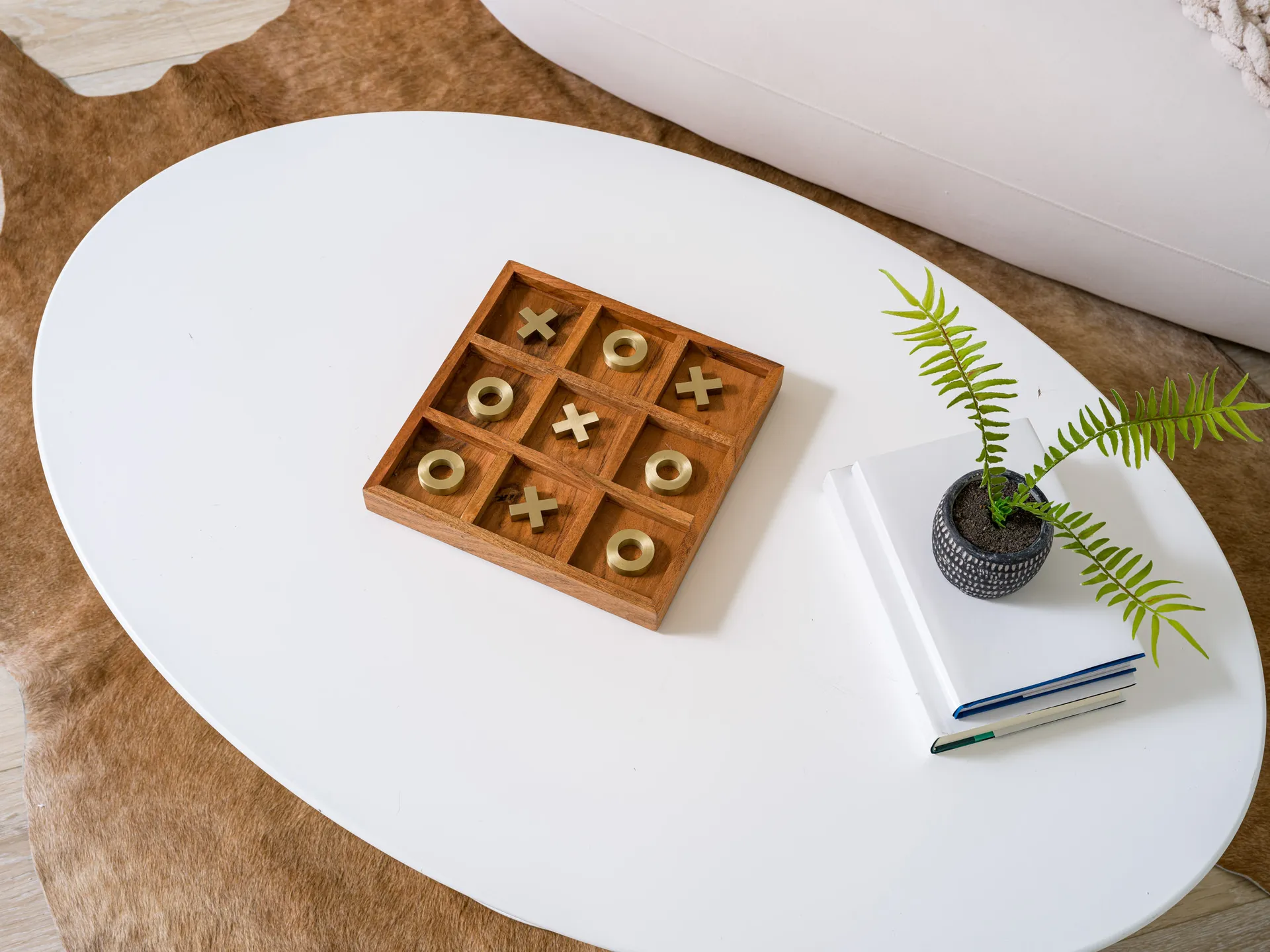 An overhead view of a white coffee table with a wooden tic-tac-toe board featuring gold X and O pieces, next to a small potted plant on stacked books.