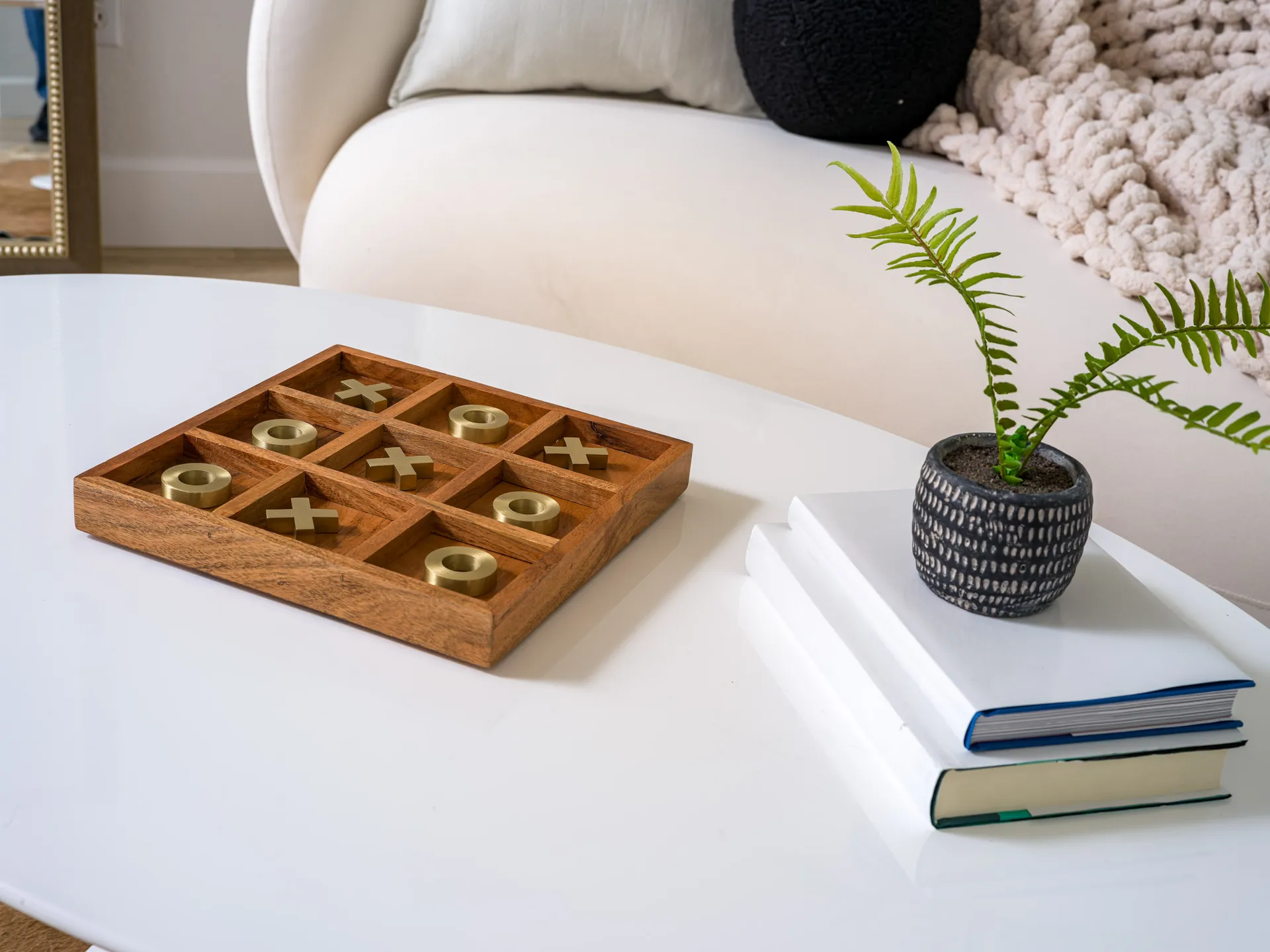 A wooden tic-tac-toe set with gold pieces on a white coffee table, next to a small potted plant and books.