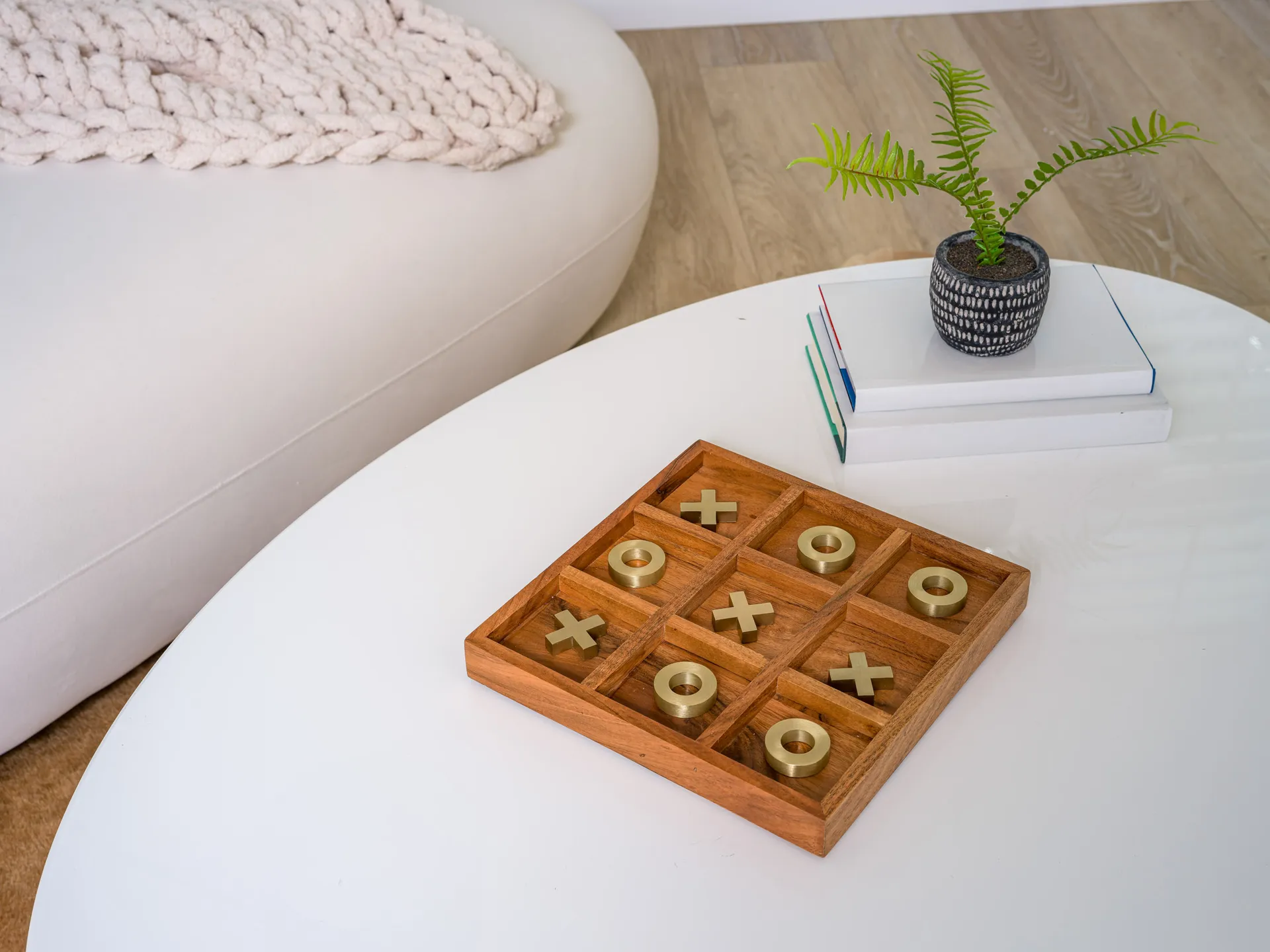 A wooden tic-tac-toe board with gold X and O pieces on a white coffee table, alongside a small potted plant and stacked books.