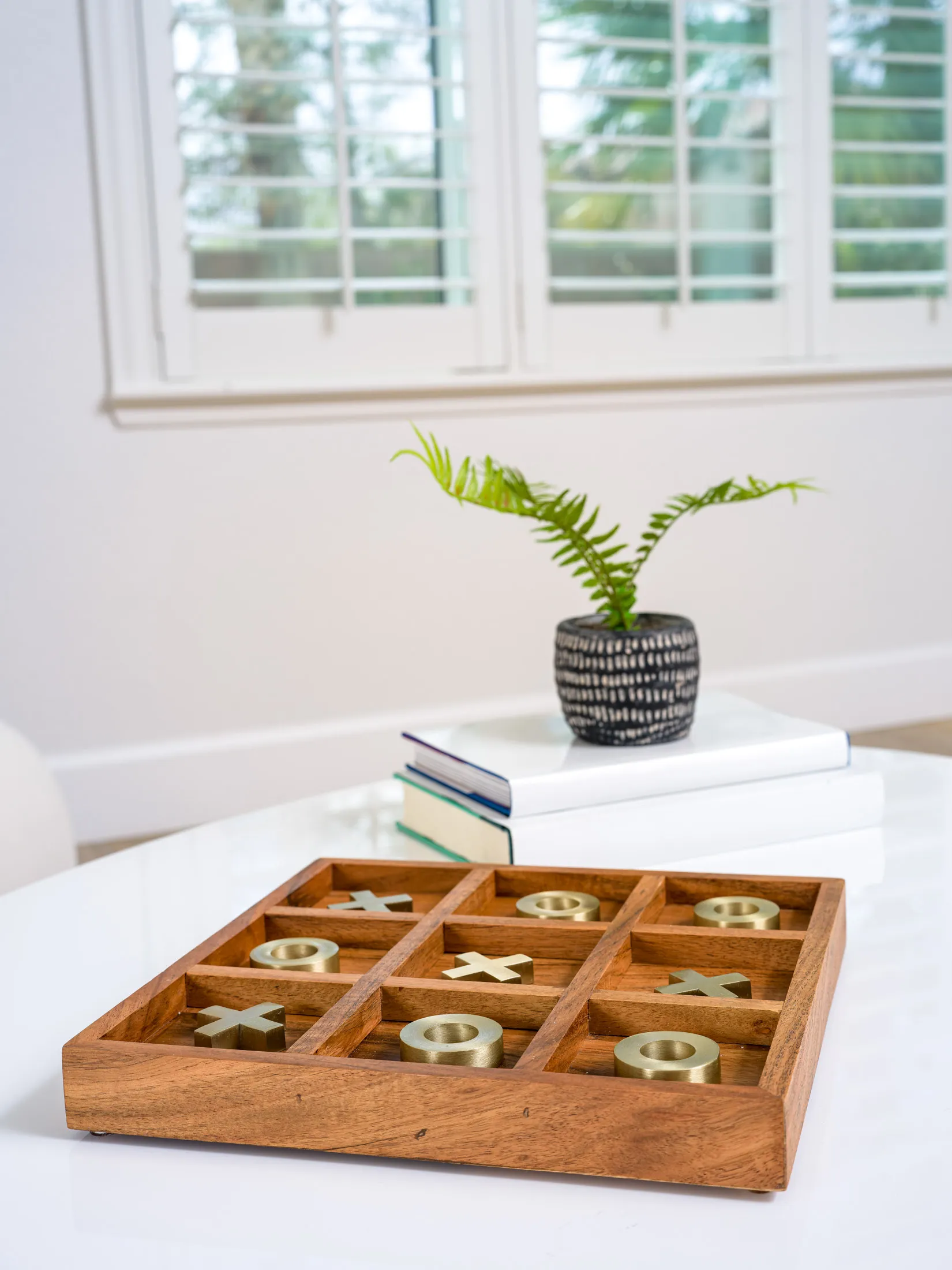 A wooden tic-tac-toe board with gold X and O pieces on a white tabletop, next to a small plant on stacked books near a window.