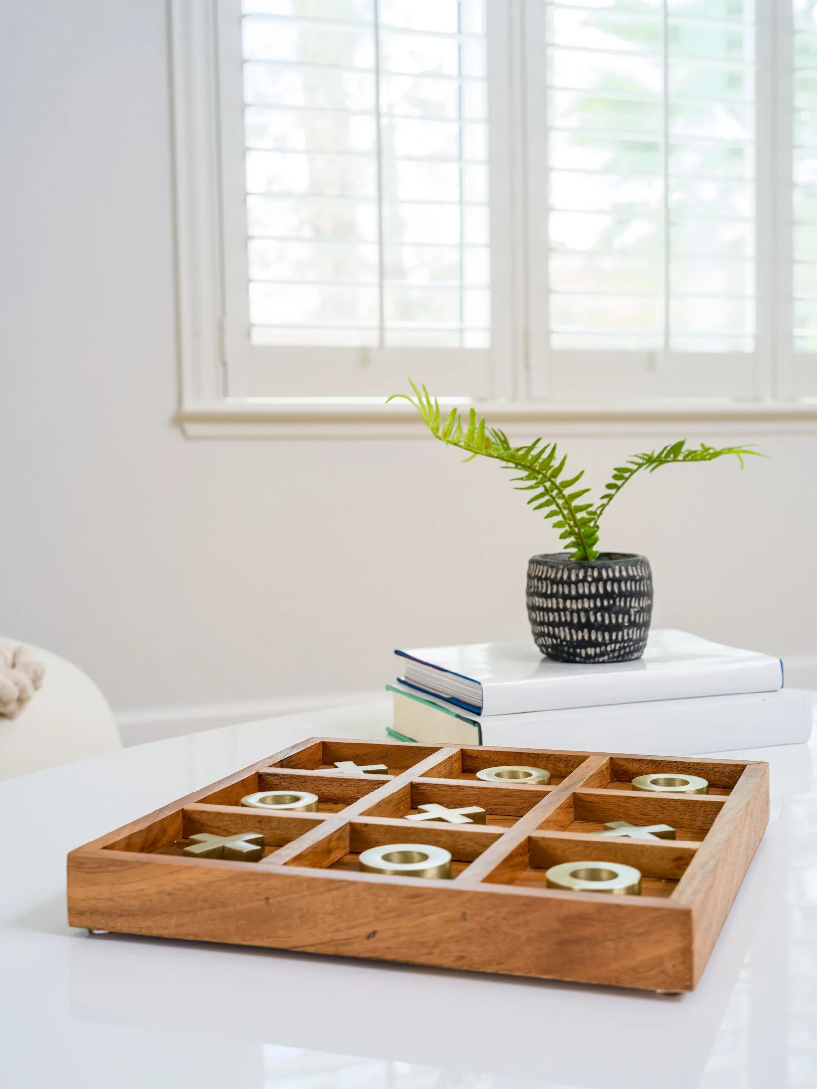 A wooden tic-tac-toe board with gold X and O pieces on a white table, near a small potted plant and stacked books.