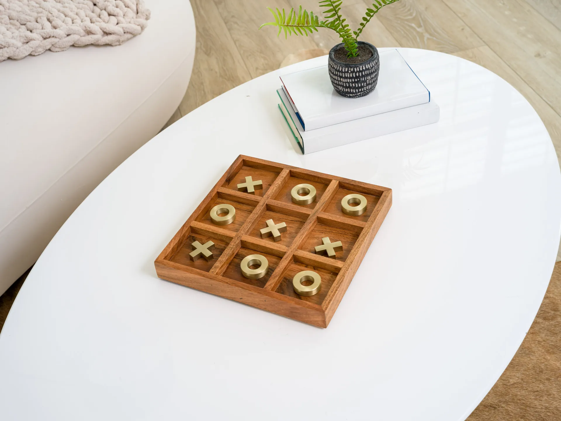A wooden tic-tac-toe board with gold X and O pieces on a white coffee table, near a small plant and stacked books.