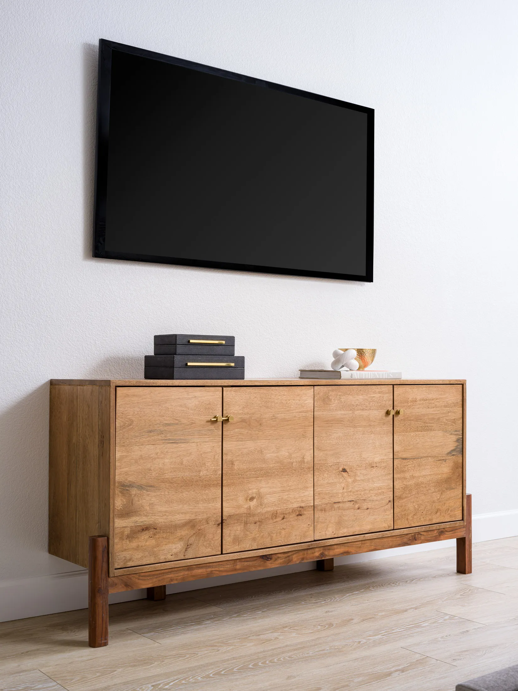 A wooden sideboard with gold hardware beneath a mounted TV, topped by black boxes, a gold bowl, and a white knot sculpture.