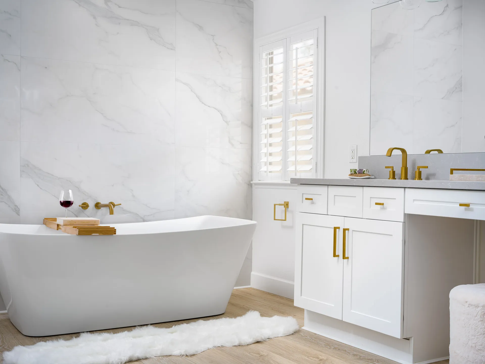 A white freestanding bathtub with a bamboo tray, gold fixtures, and white cabinets in a bright bathroom.