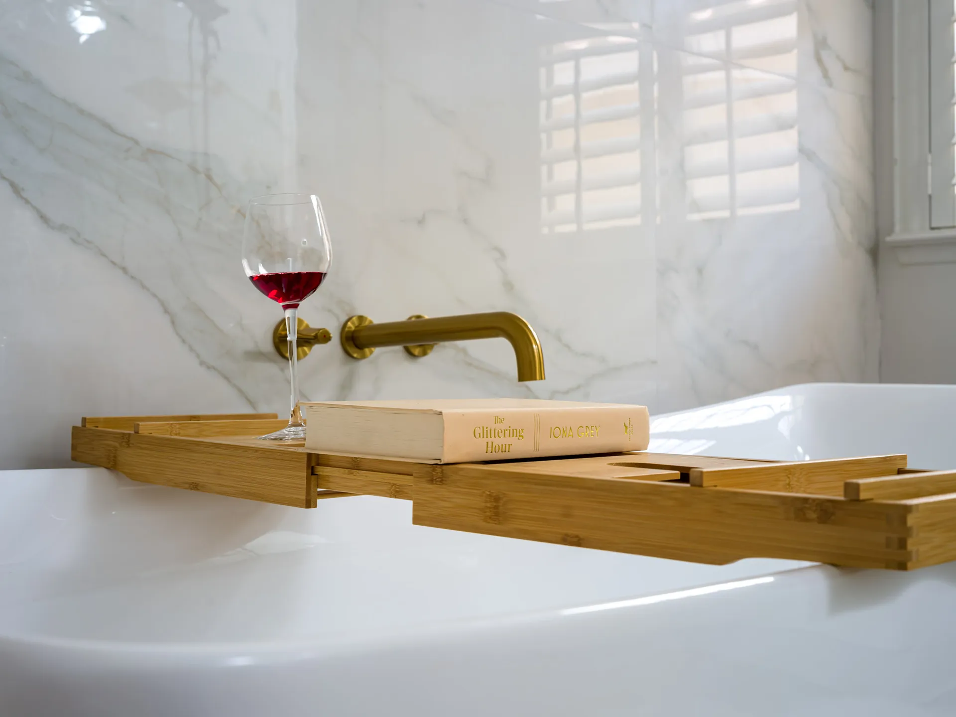 A white bathtub with gold hardware and a bamboo tray holding a glass of red wine and a book.