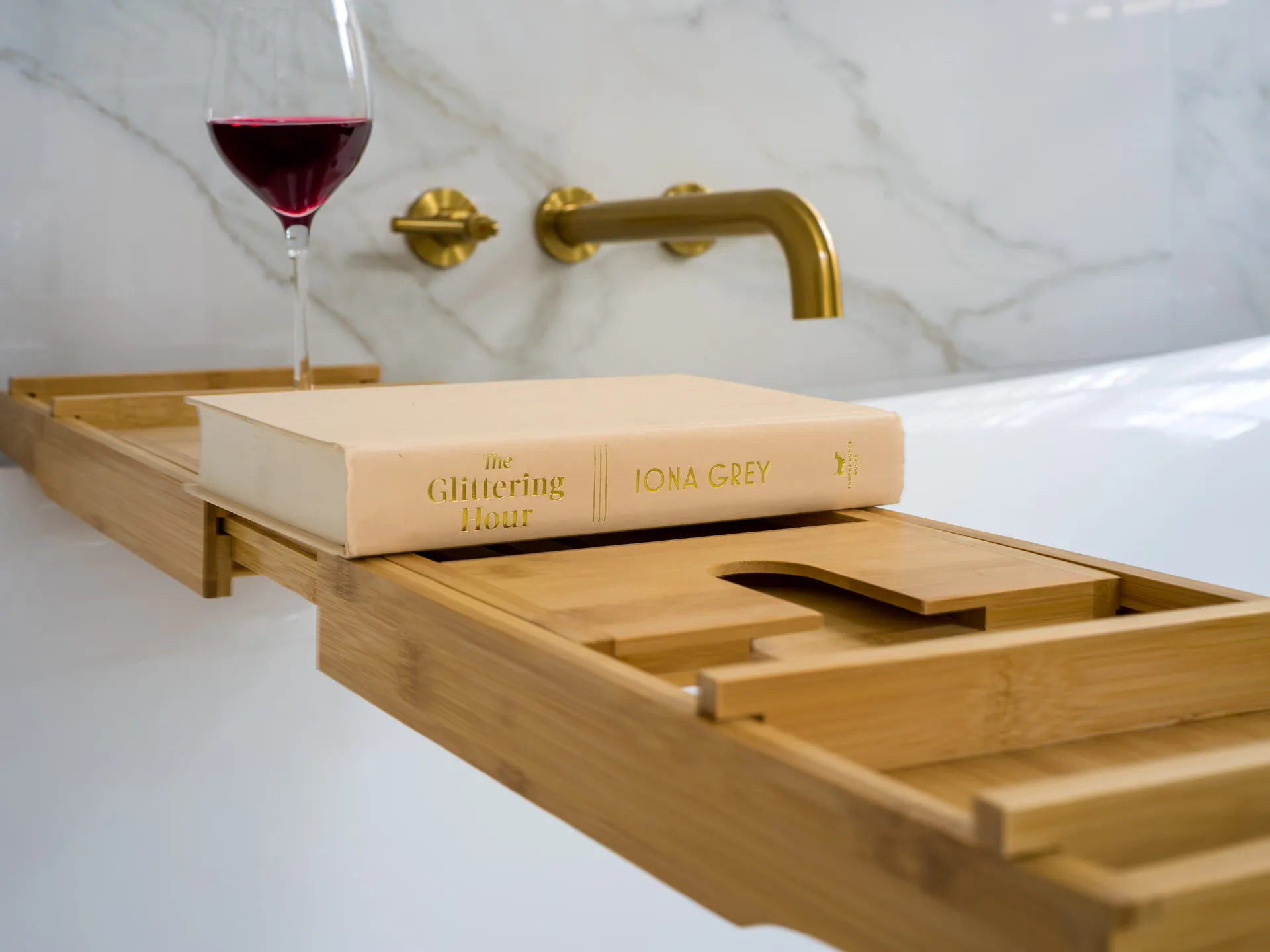 A bamboo bathtub tray holds a hardcover book and a glass of red wine, set against a marble wall with gold fixtures