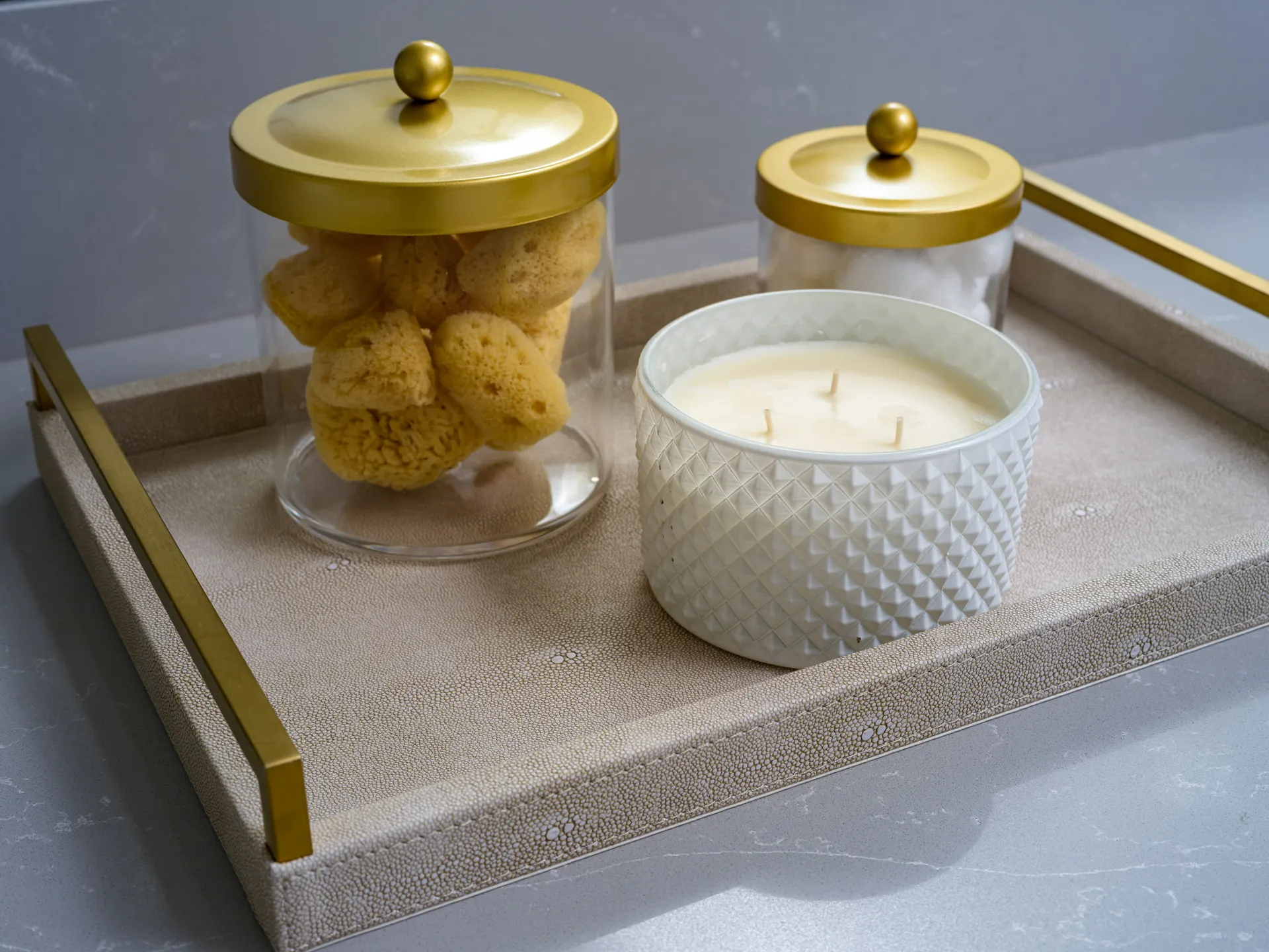 A beige tray with gold handles holds a white textured candle and two glass jars with gold lids, one containing natural sponges and the other cotton balls.