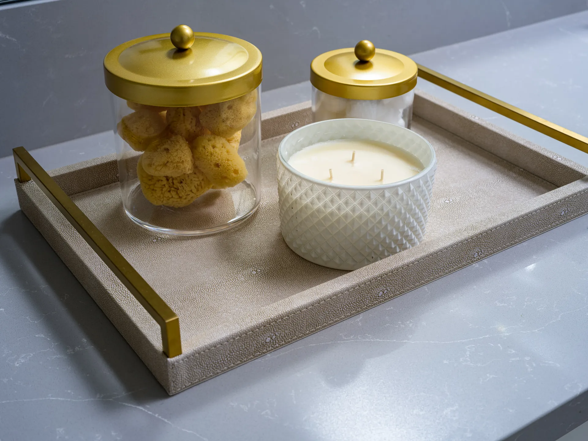 A neutral tray on a gray countertop, holding gold-lidded jars of sponges and cotton balls along with a textured white candle.