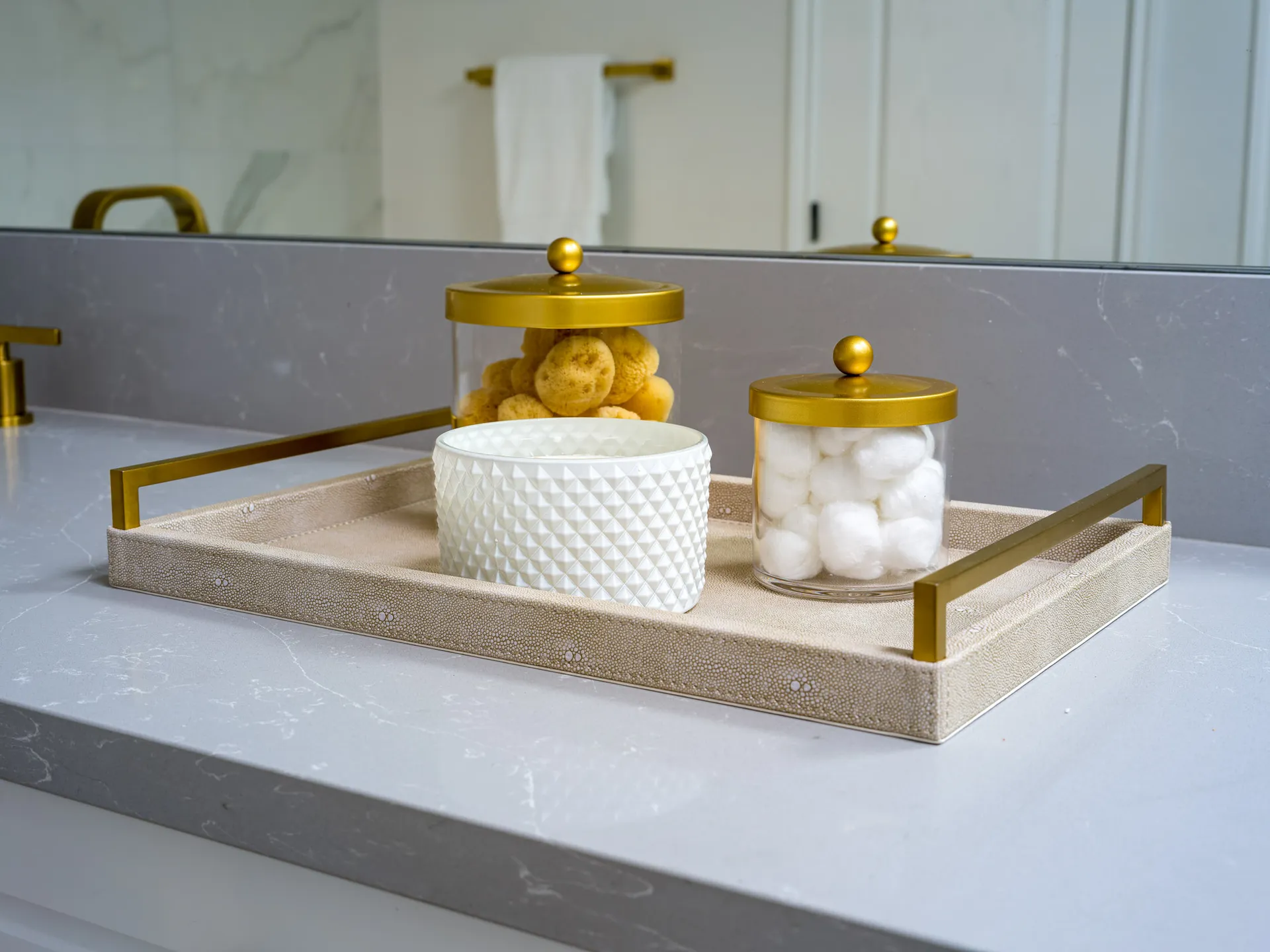A bathroom countertop with a neutral tray holding two glass jars with gold lids—one filled with sponges, the other with cotton balls—and a textured white candle.