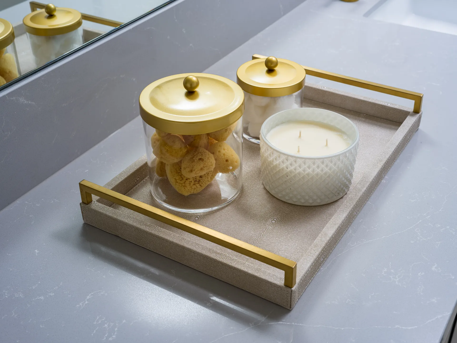 A neutral tray on a gray countertop displaying gold-lidded jars filled with sponges and cotton balls, alongside a textured white candle.