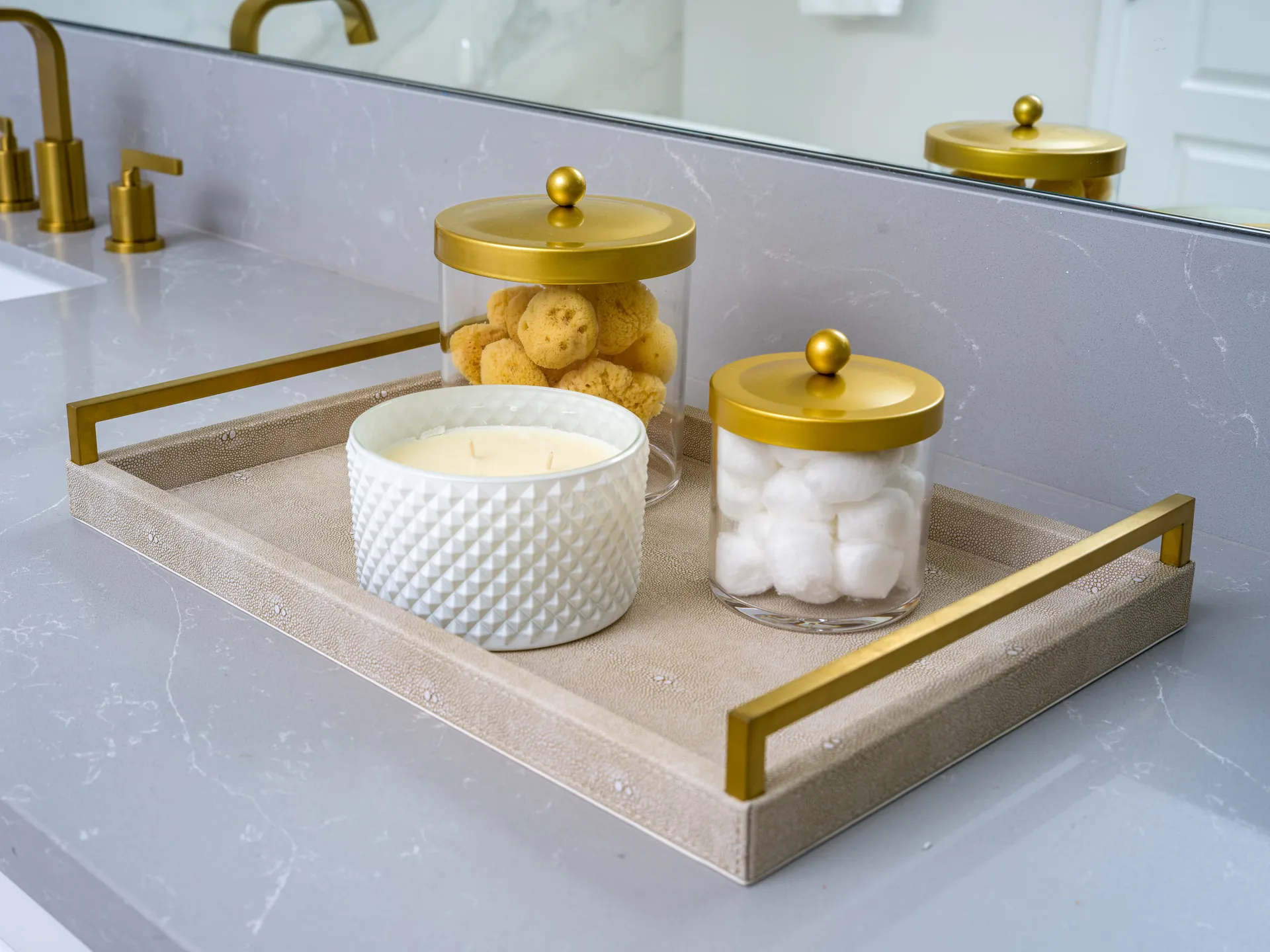 A gold-handled tray on a bathroom counter with glass canisters containing sponges and cotton balls, along with a textured candle holder.