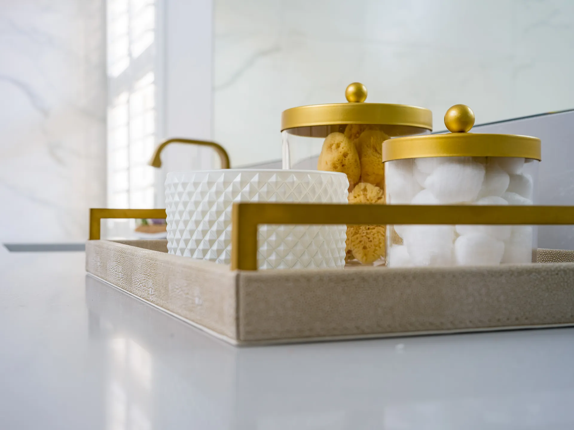 A neutral tray on a bathroom counter holding gold-lidded jars with sponges and cotton balls, alongside a textured white container.