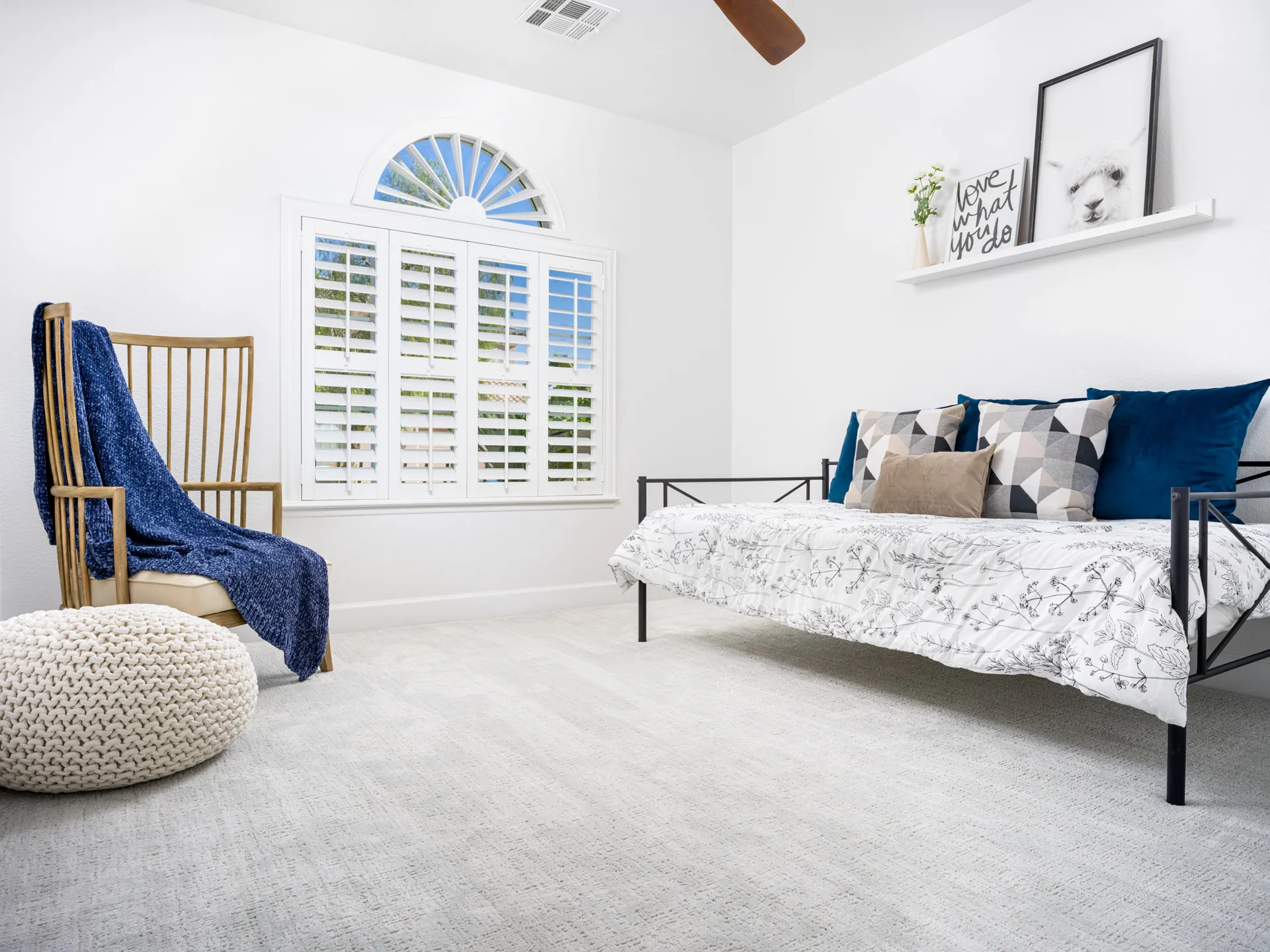 A bright bedroom featuring a black metal daybed with patterned pillows, a wooden accent chair draped in a navy throw, and a chunky knit pouf on a soft gray carpet.