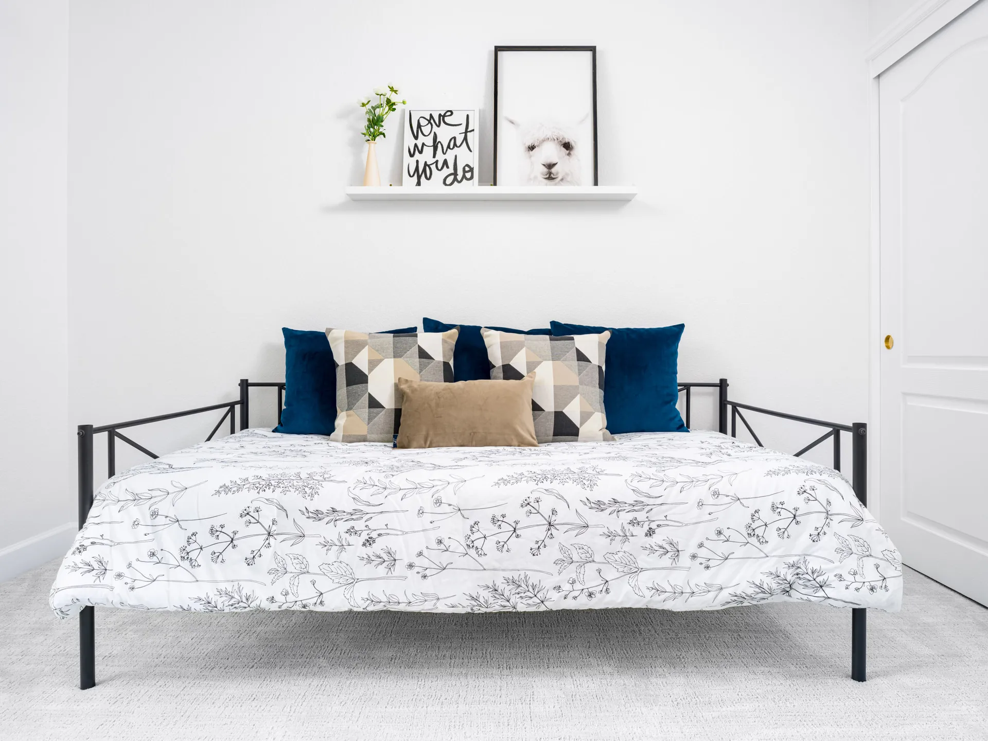 A black metal daybed dressed in a floral duvet and layered throw pillows in shades of blue and beige. A floating shelf above holds a whimsical animal print and motivational art.