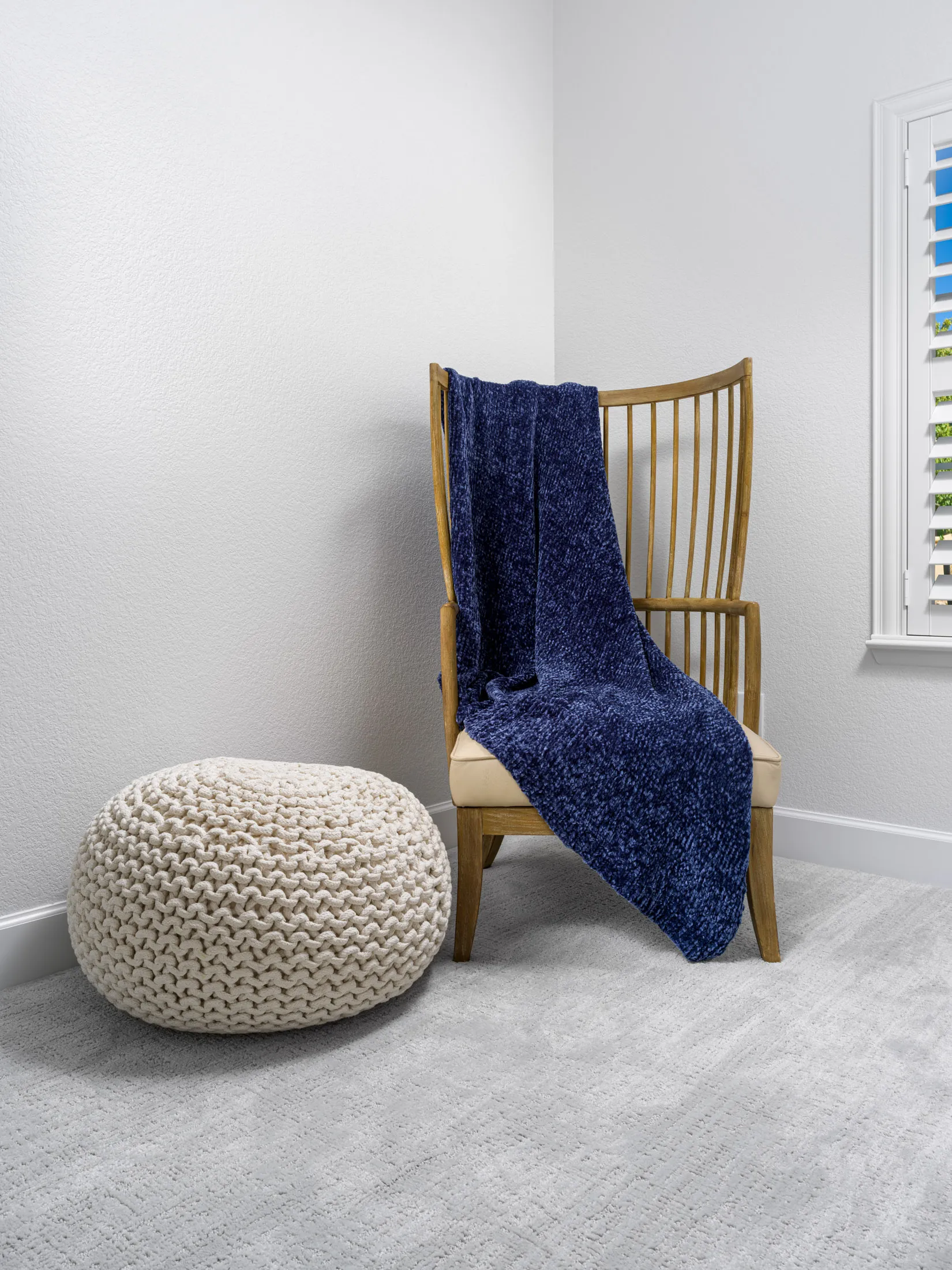 A wooden lounge chair draped with a navy throw sits next to a chunky knit pouf on a soft gray carpet. The white walls provide a clean, minimal backdrop.