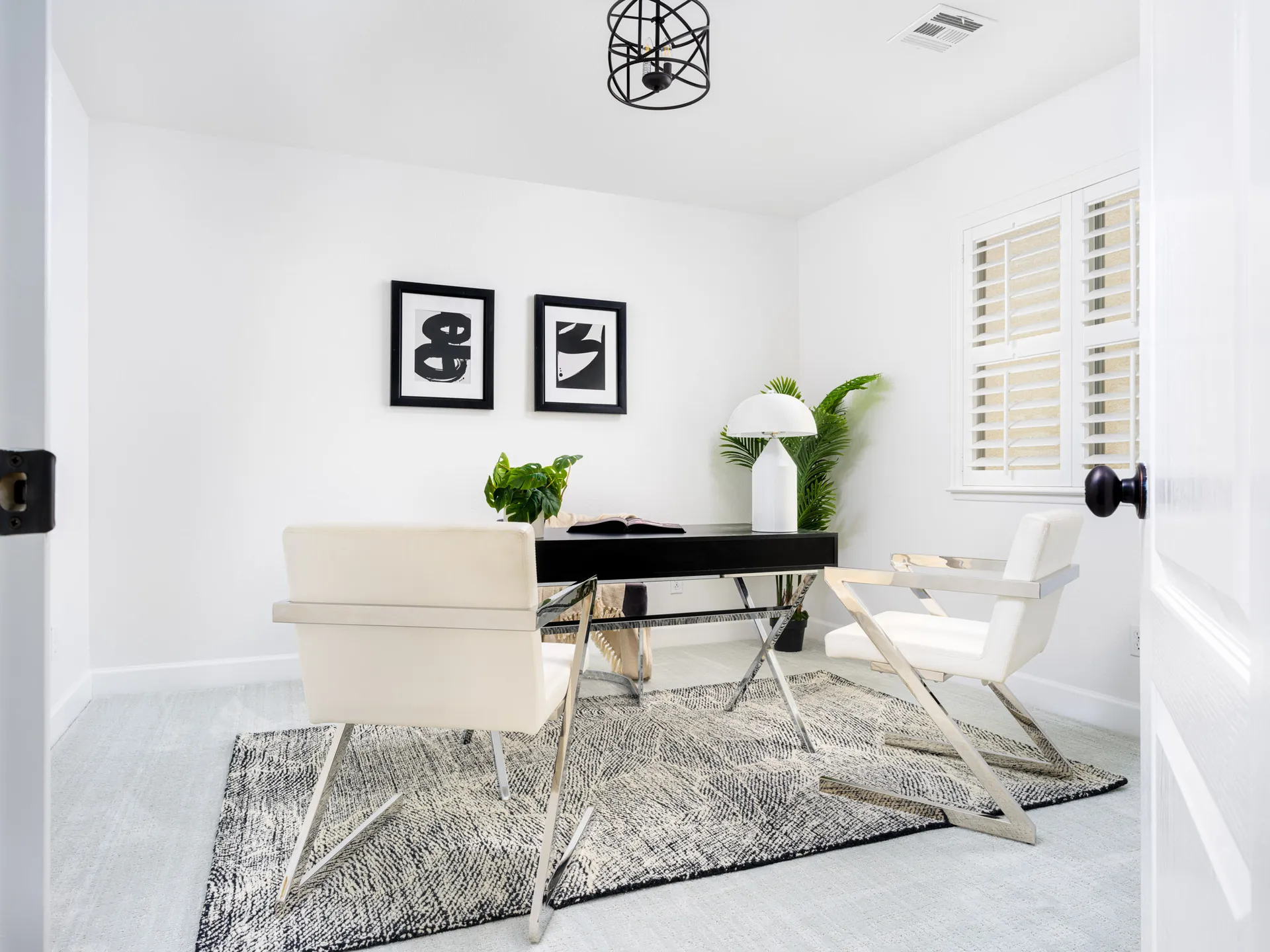 A compact office with a black desk, white chairs featuring chrome frames, and abstract black-and-white artwork on the wall. A gray-toned rug and leafy plant complete the modern look.