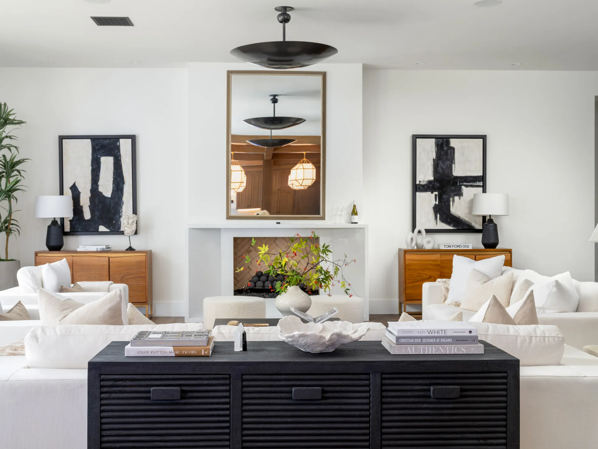 A bright living room featuring a black console table with designer books, a white sofa, a fireplace, and matching wooden sideboards with abstract art.