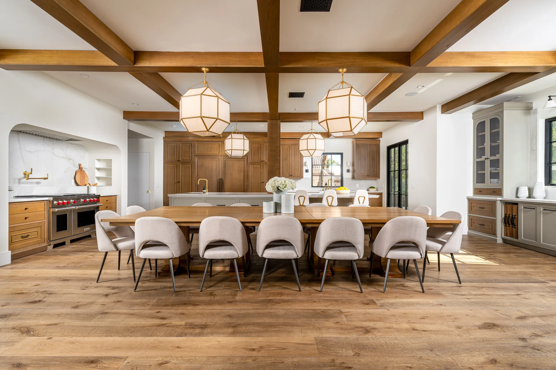 A spacious dining area with a large wooden table, upholstered chairs, exposed wood beams, and an open-concept kitchen with natural light.