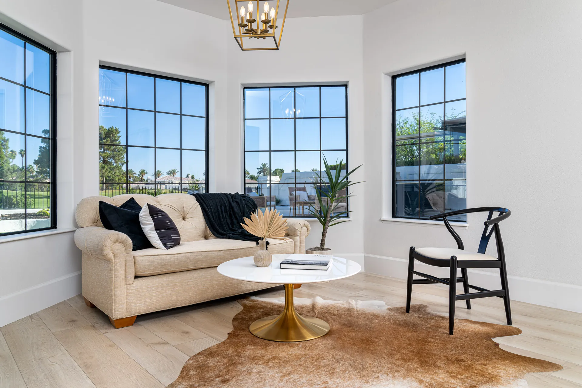 Cozy living room with a beige sofa, a round white coffee table with a gold base, a black armchair, and a cowhide rug. Large windows with black frames and a pendant light hanging from the ceiling create a bright and inviting space.