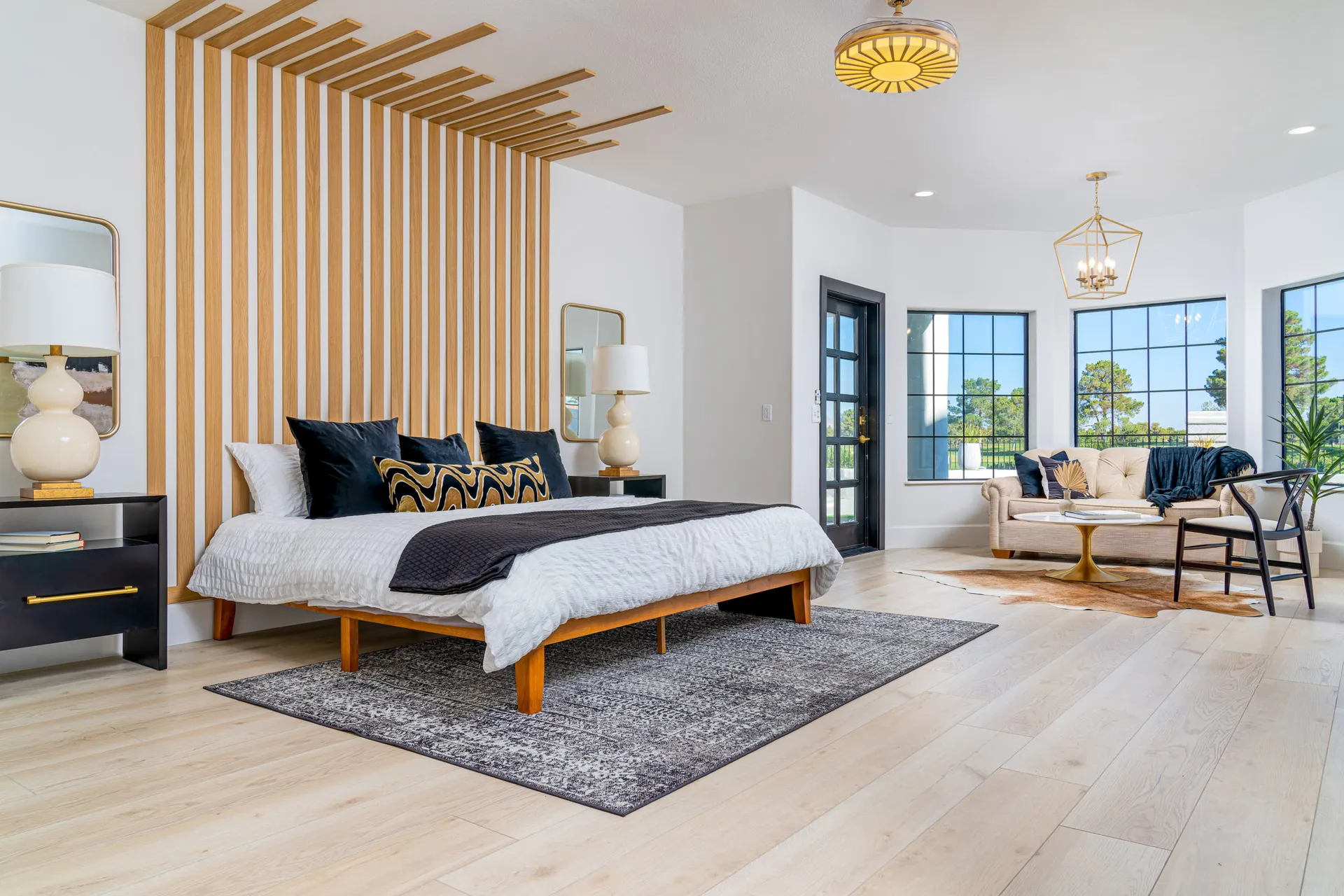 Bedroom with a wooden slat accent wall, a bed with white linens, black pillows, and a black throw blanket. There are two black nightstands with gold accents, and a beige rug on the floor. The room has a modern and cozy design, creating a comfortable and stylish sleeping space.