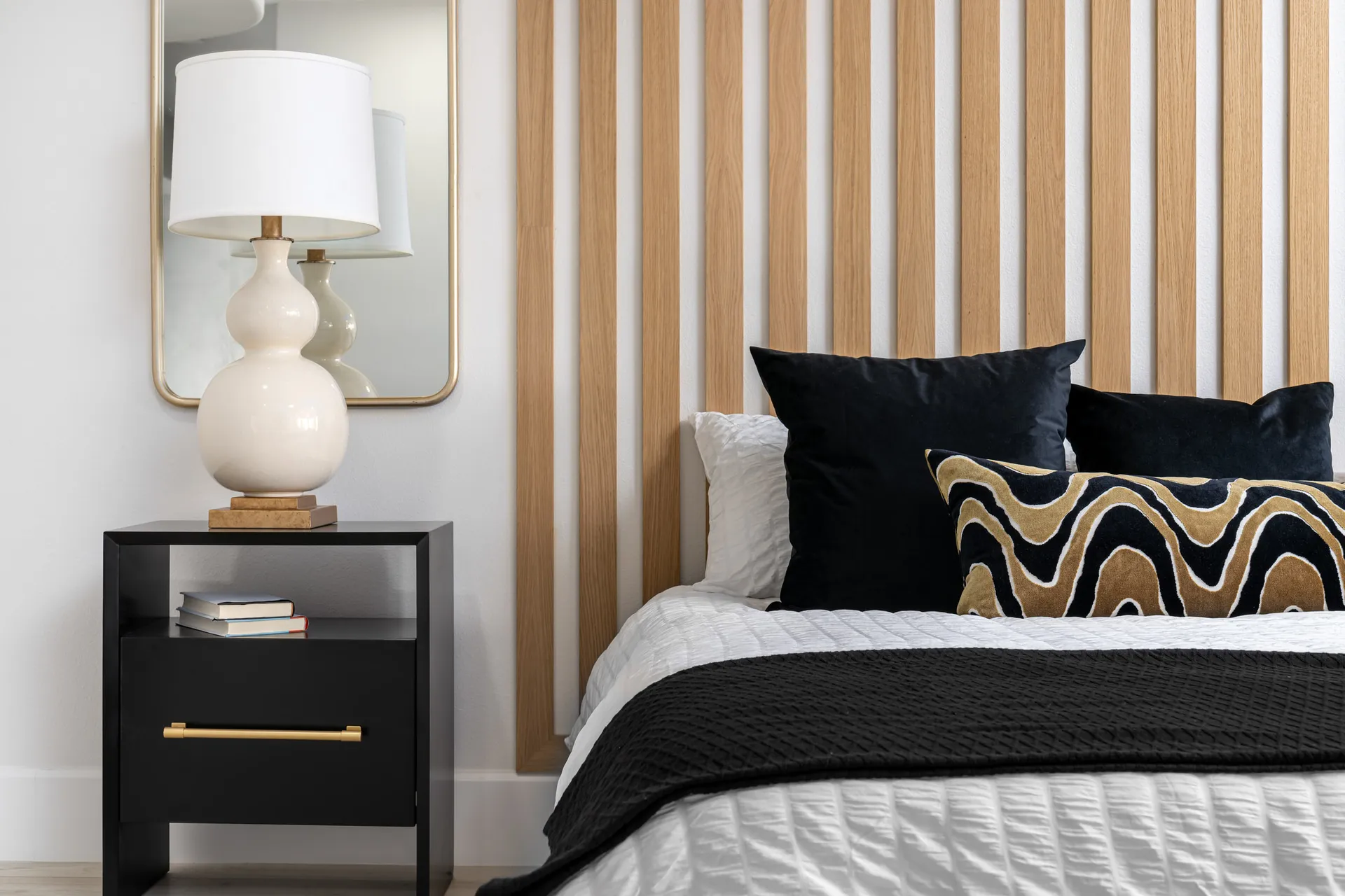 Close-up of a bedroom with a wooden slat accent wall, a bed with white linens, black pillows, and a black throw blanket. There is a black nightstand with gold accents and a table lamp, creating a modern and cozy sleeping space.