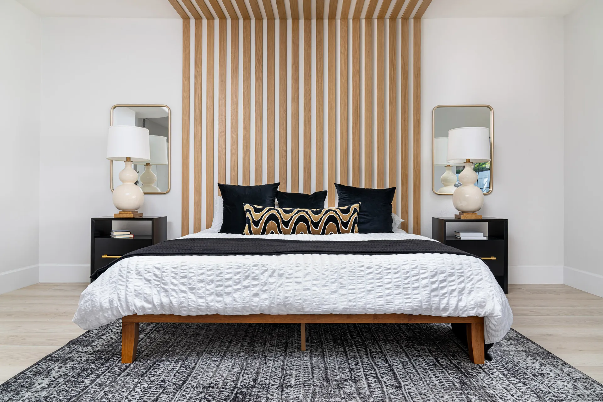 Bedroom with a wooden slat accent wall, a bed with white linens, black pillows, and a black throw blanket. There are two black nightstands with gold accents, and a beige rug on the floor. The room has a modern and cozy design, creating a comfortable and stylish sleeping space.