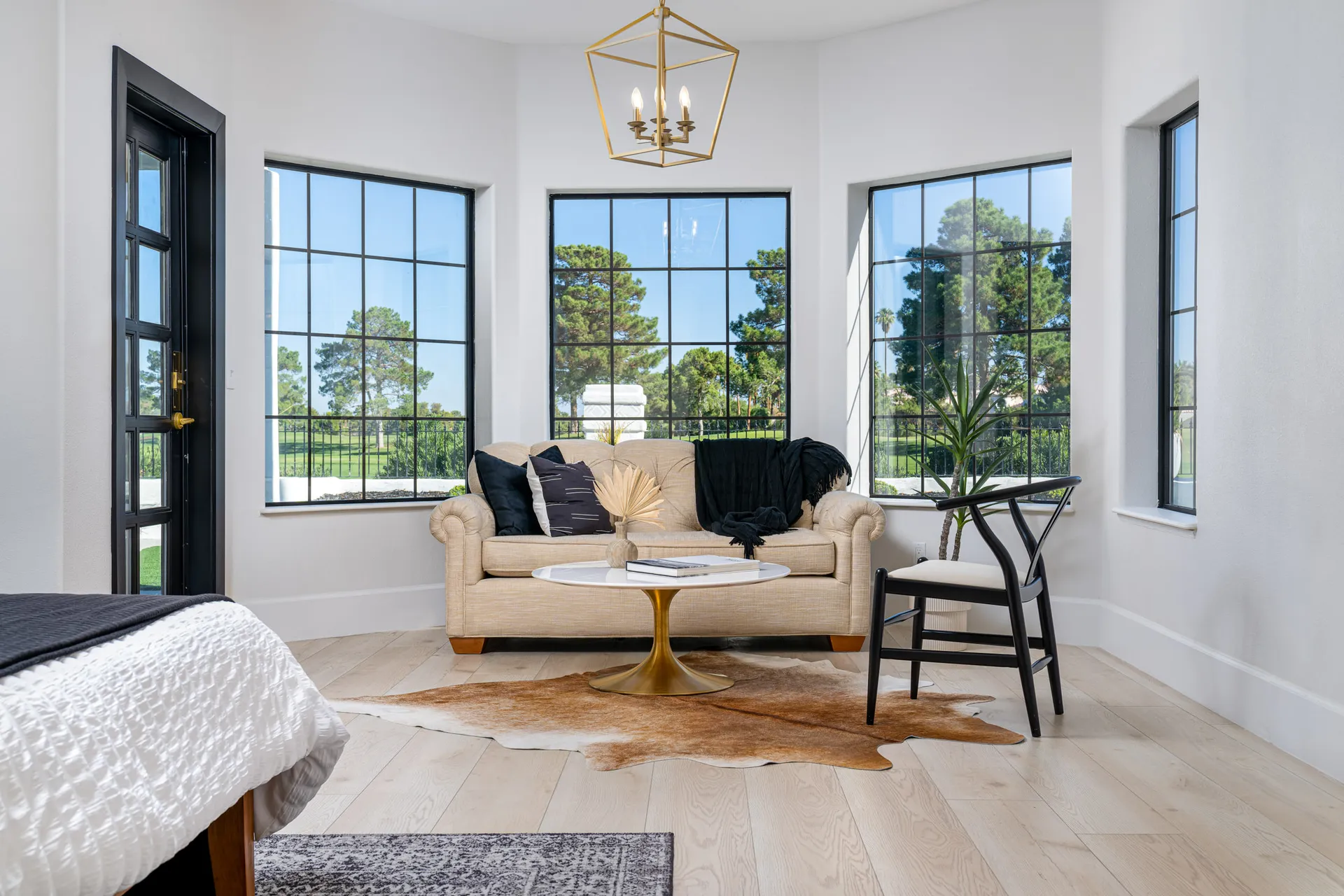 A beige sofa with black accent pillows sits near large windows overlooking a golf course, with a gold pendant chandelier overhead.