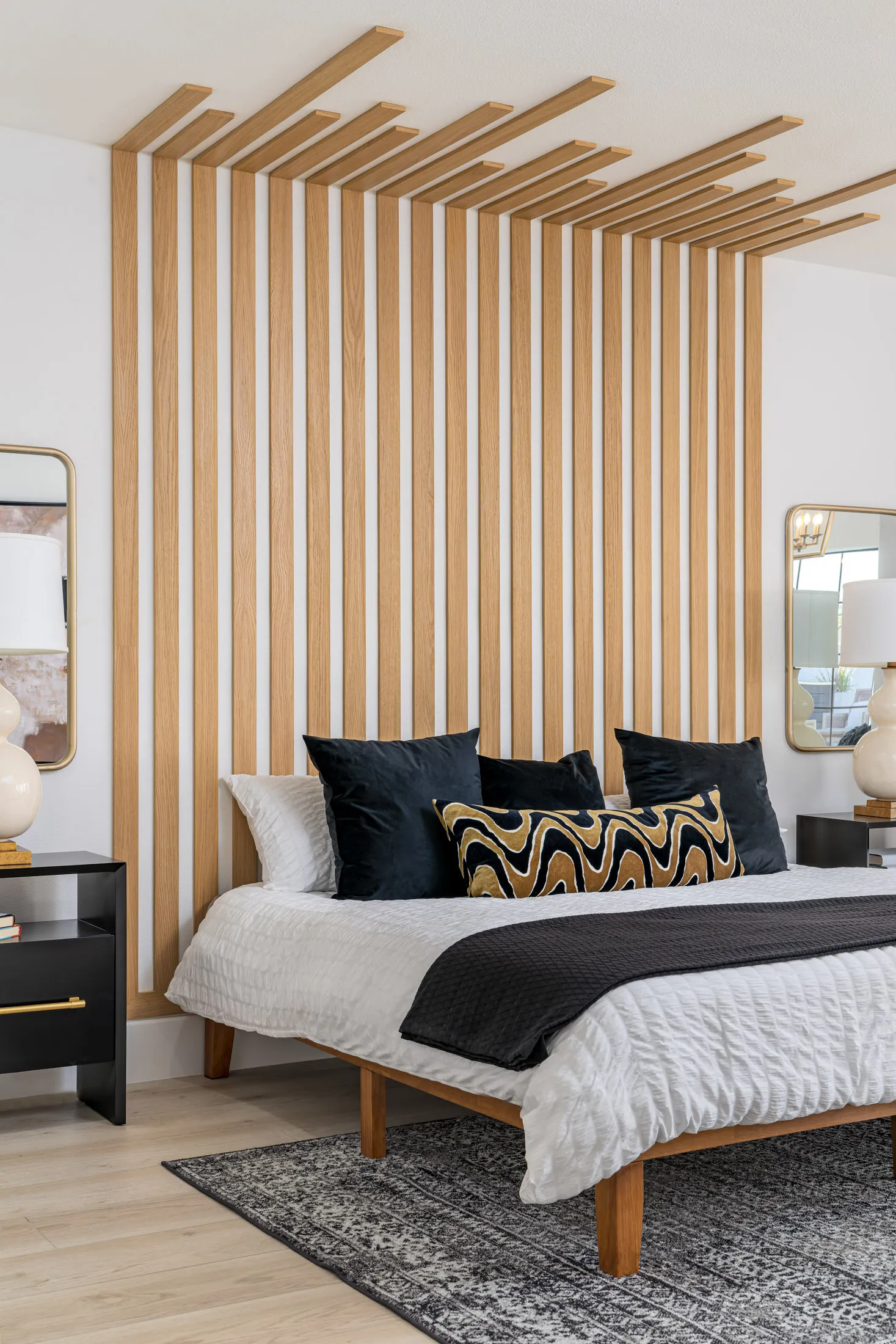 Bedroom with a wooden slat accent wall, a bed with white linens, black pillows, and a black throw blanket. There are two black nightstands with gold accents, and a beige rug on the floor. The room has a modern and cozy design, creating a comfortable and stylish sleeping space.
