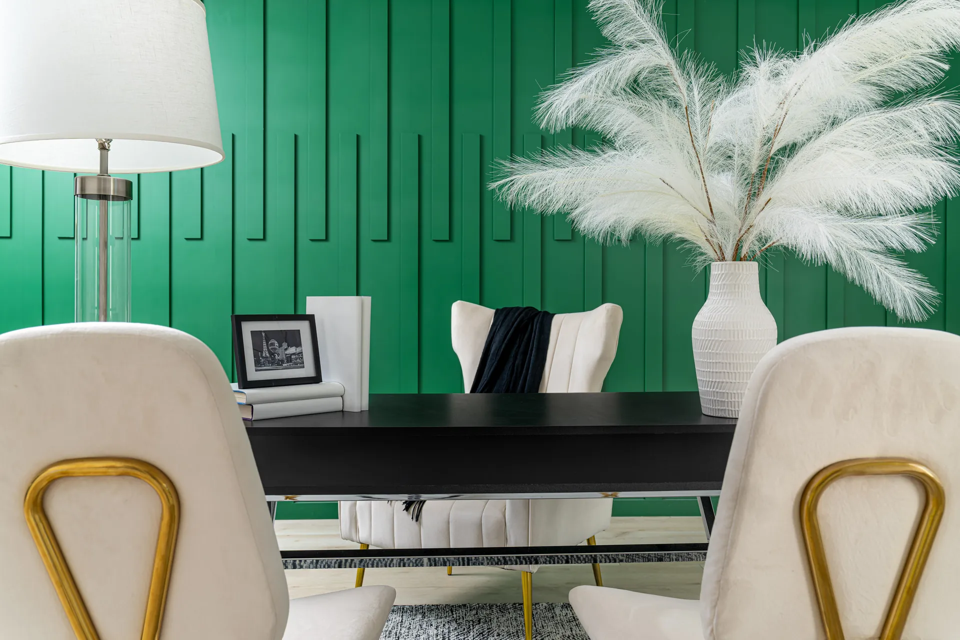 Dining table with a black top and gold chairs, set against a green wall with vertical lines. There are decorative items like a vase with pampas grass and a table lamp. The room has a modern and elegant design, creating a stylish dining space.
