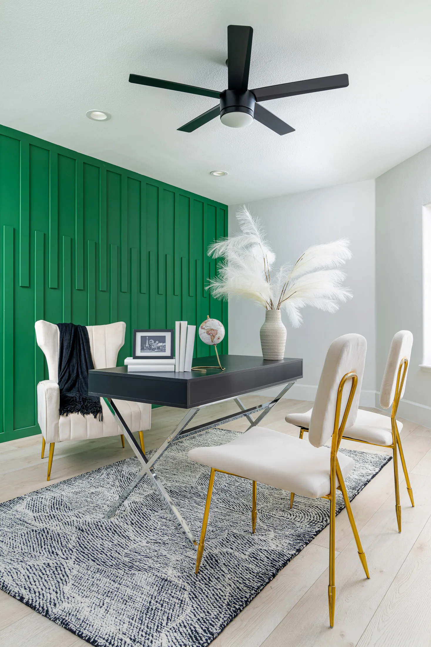 Home office with a black desk, a large green cabinet, and a white chair. There are decorative items like a vase with pampas grass, a framed quote, and a black ceiling fan. The room has a neutral color palette with a pop of green from the cabinet, creating a stylish and functional workspace.