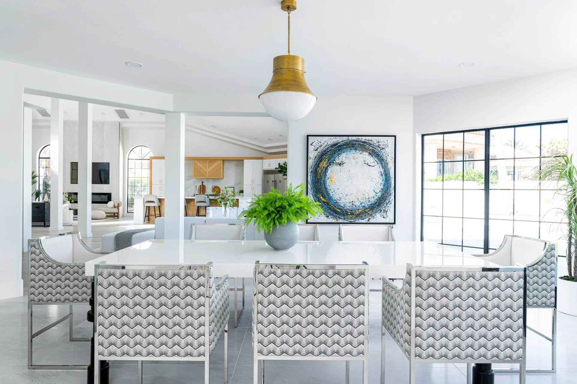 Dining area with a white marble table, patterned chairs, and a large abstract painting on the wall. The room has a gold pendant light, and the background includes a view of the kitchen and living area, creating an elegant and cohesive living space.
