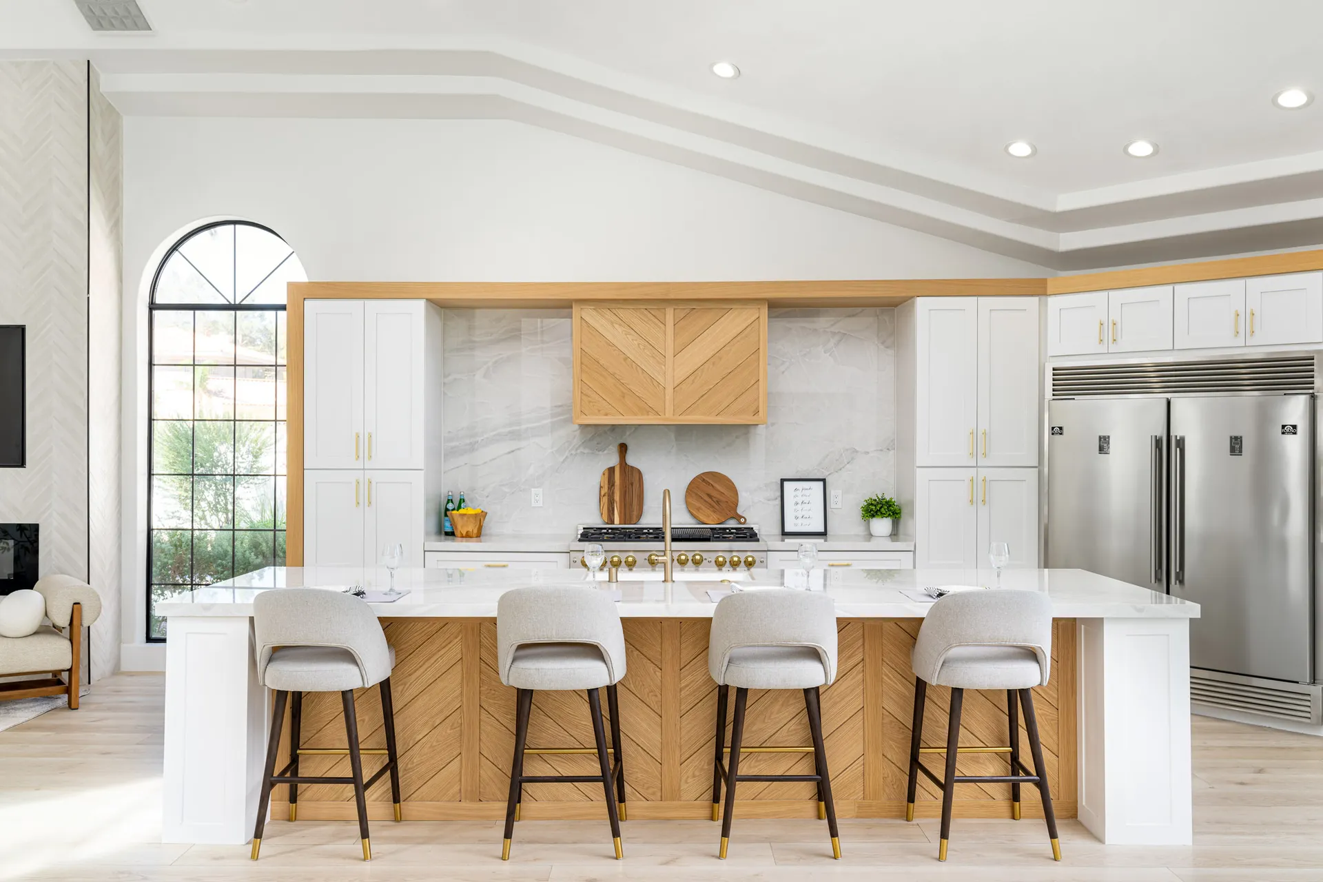Kitchen with a large white marble island, wooden bar stools, and white cabinets with gold handles. The backsplash is white marble, and there are decorative items like a wooden cutting board, a framed quote, and a plant, creating a luxurious and elegant kitchen space.