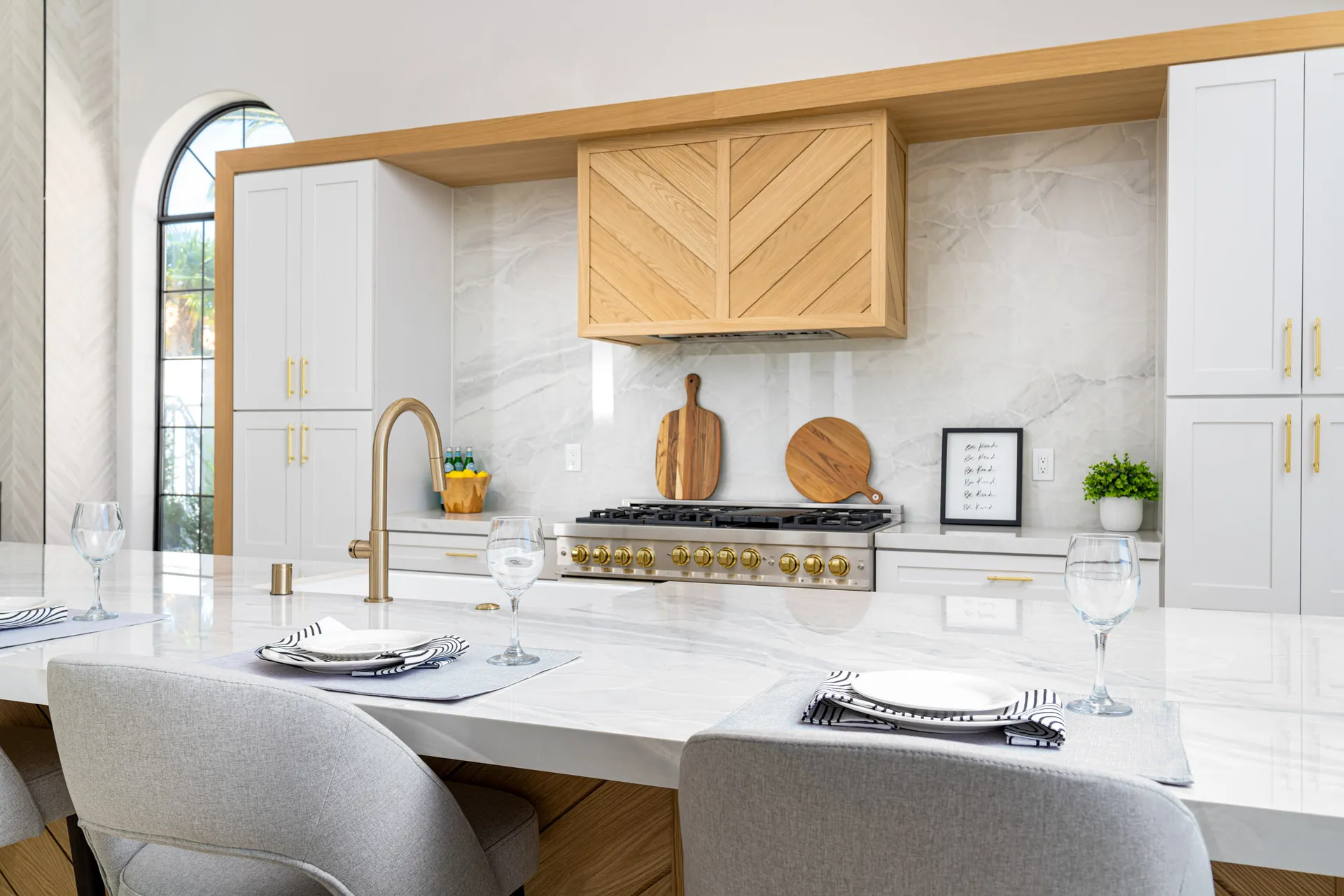 Kitchen with a white marble countertop, gold-accented gas stove, and white cabinets with gold handles. The backsplash is white marble, and there are decorative items like a wooden cutting board, a framed quote, and a plant, creating a luxurious and elegant kitchen space.